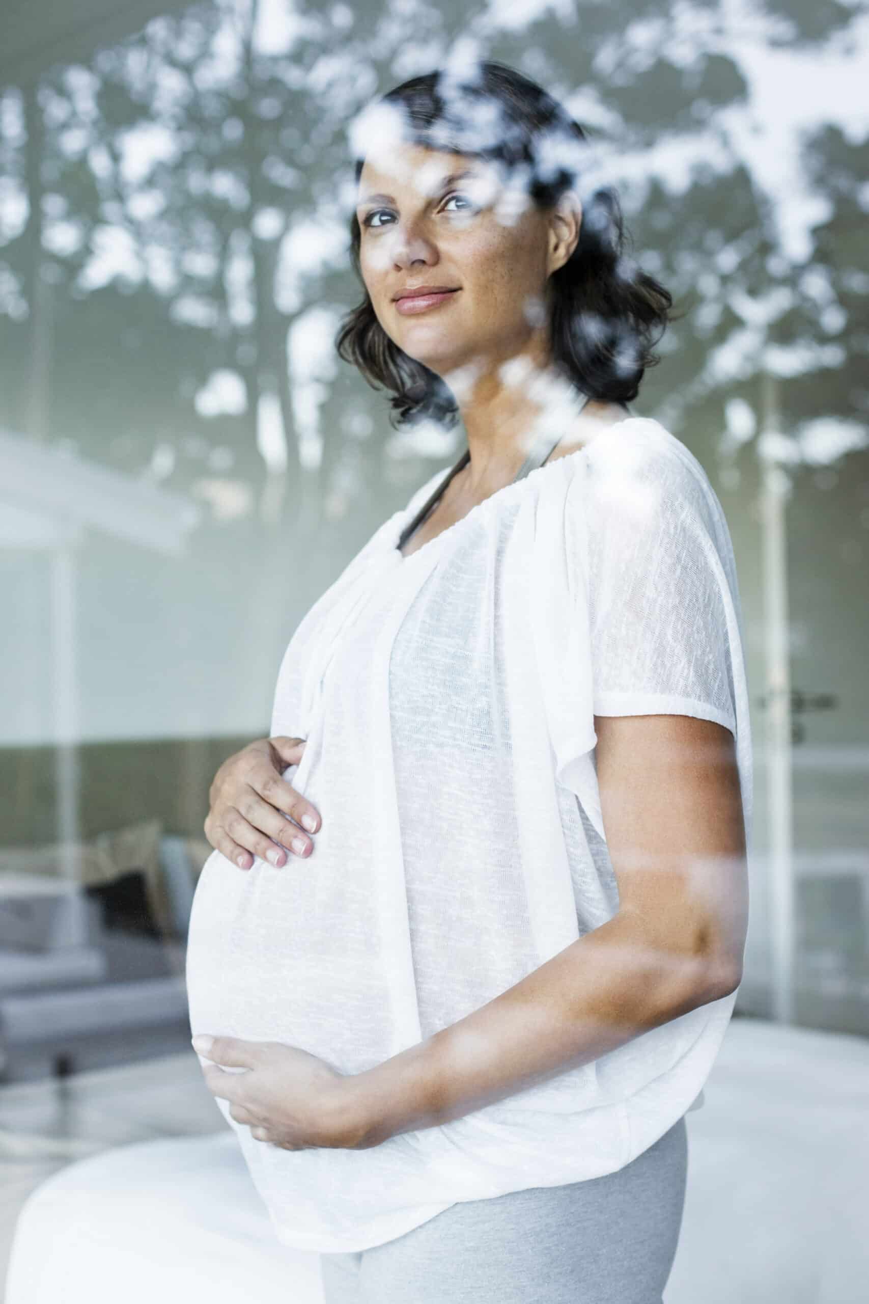 Eine schwangere Frau in einer weißen Bluse steht drinnen und blickt aus einem Fenster mit spiegelnder Oberfläche. Sie hält sanft ihren Bauch und sieht nachdenklich und gelassen aus. © Fotografie Tomas Rodriguez