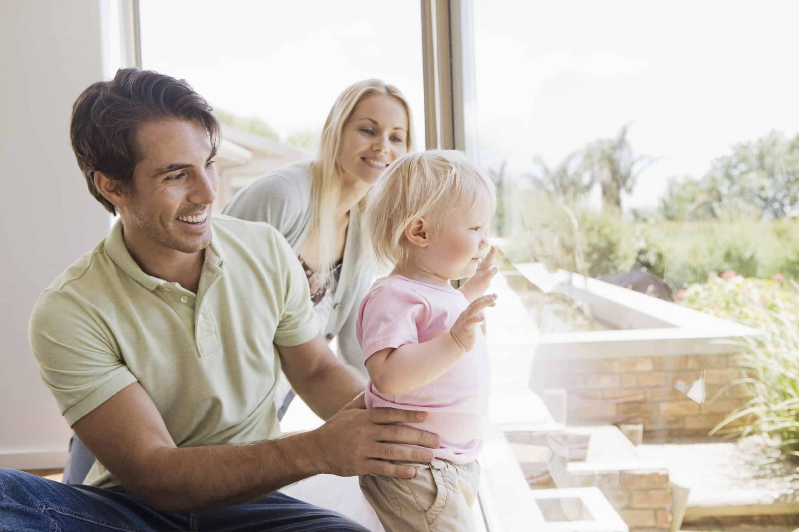 Eine glückliche Familie in einem hellen Raum: Ein Mann und eine Frau lächeln ein Kleinkind an, das dasteht und wegschaut. Die drei stehen vor einem großen Fenster, durch das das Sonnenlicht hereinströmt. © Fotografie Tomas Rodriguez