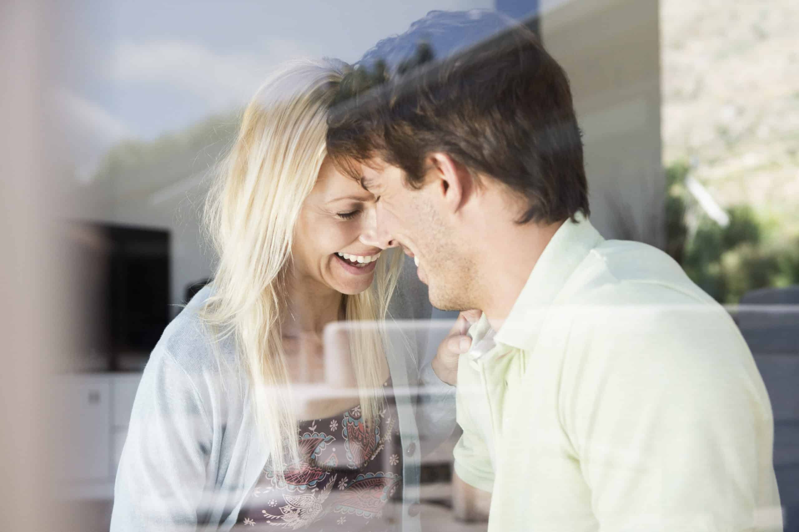 Ein fröhliches Paar berührt seine Stirn und lächelt sich durch ein Glasfenster vor einer hellen, sonnigen Kulisse an. © Fotografie Tomas Rodriguez