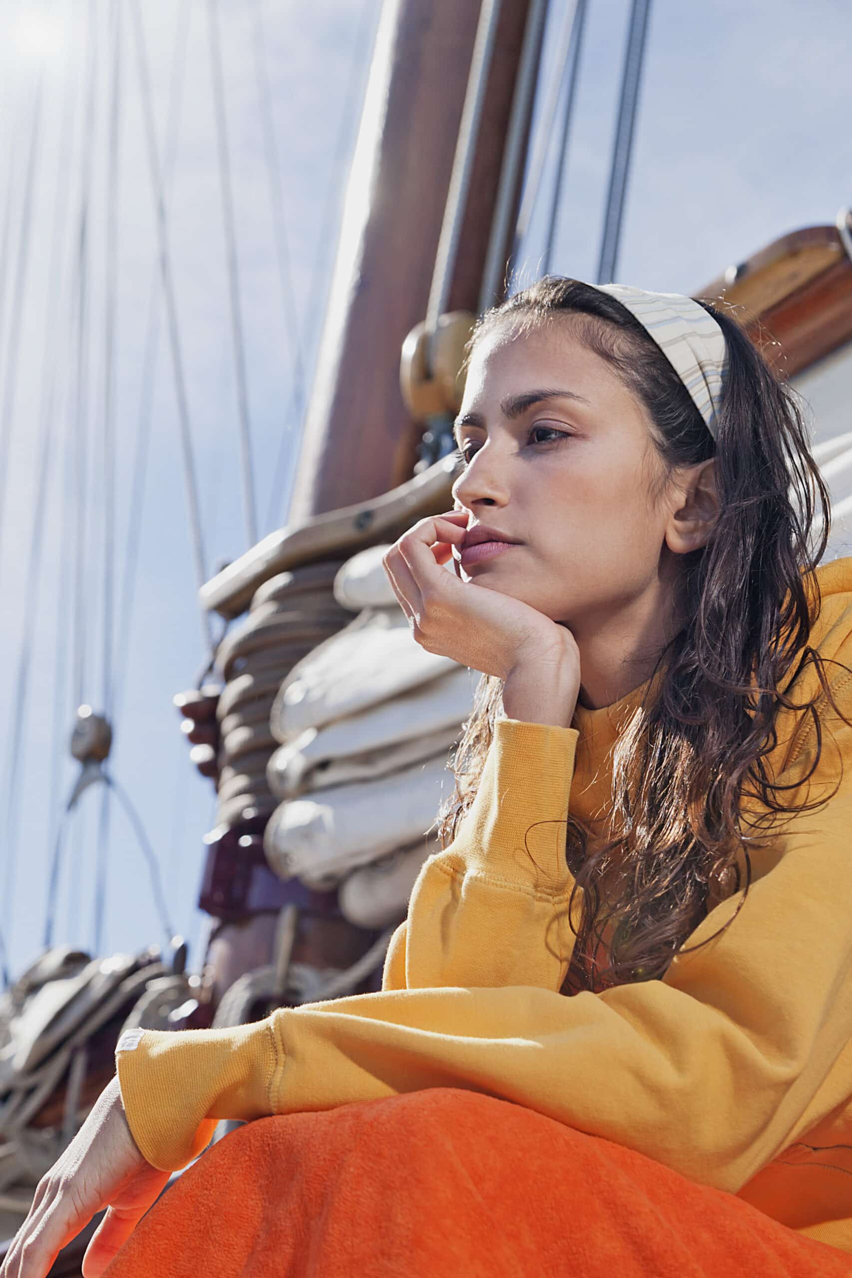 Eine Person in einem gelben Sweatshirt und einem weißen Stirnband sitzt auf einem Segelboot mit aufgerollten Segeln dahinter. Sie hat lange Haare, stützt ihr Kinn auf ihre Hand und blickt nachdenklich in die Ferne unter einem hellen, sonnigen Himmel. © Fotografie Tomas Rodriguez