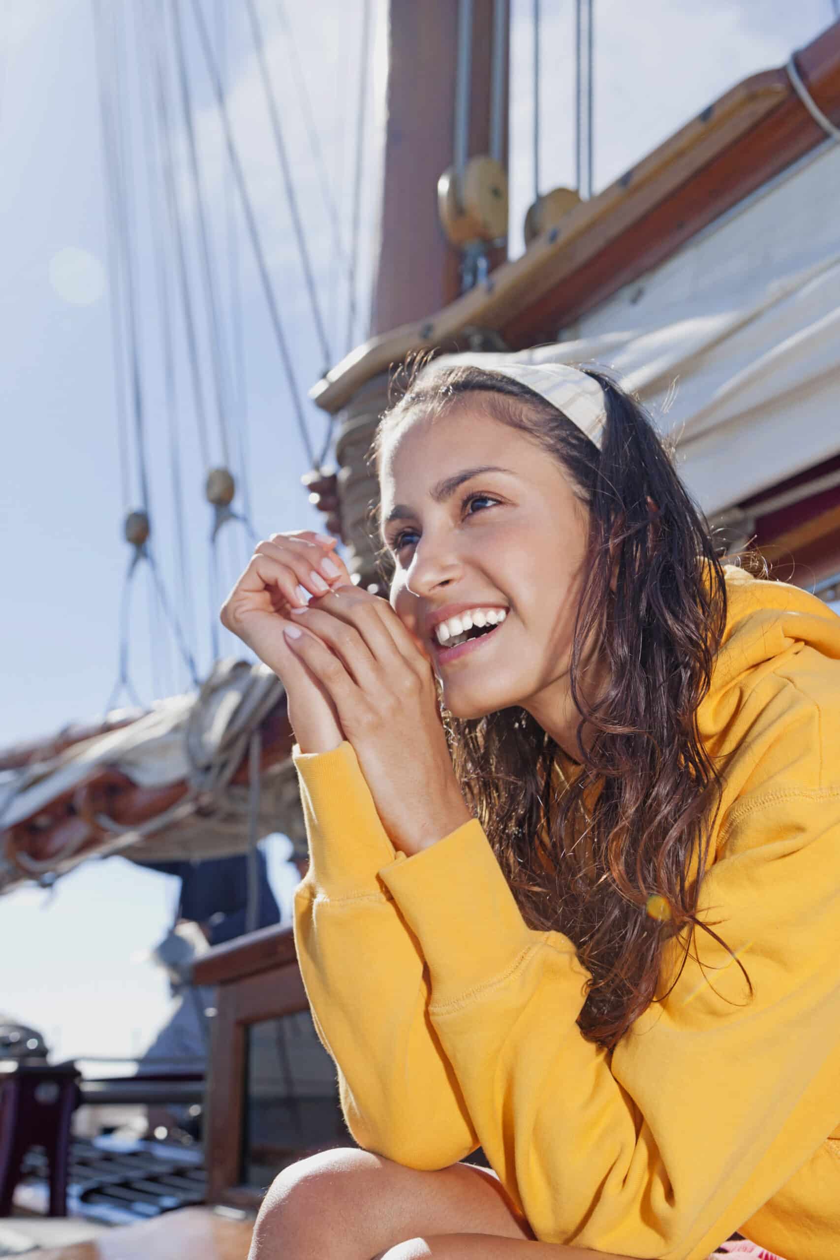 Eine junge Frau mit langen, nassen Haaren und einem gelben Kapuzenpullover sitzt strahlend auf einem Boot. Sie hält ihre Hände vors Gesicht und im Hintergrund sind Segel und Takelage vor einem klaren, sonnigen Himmel zu sehen. © Fotografie Tomas Rodriguez