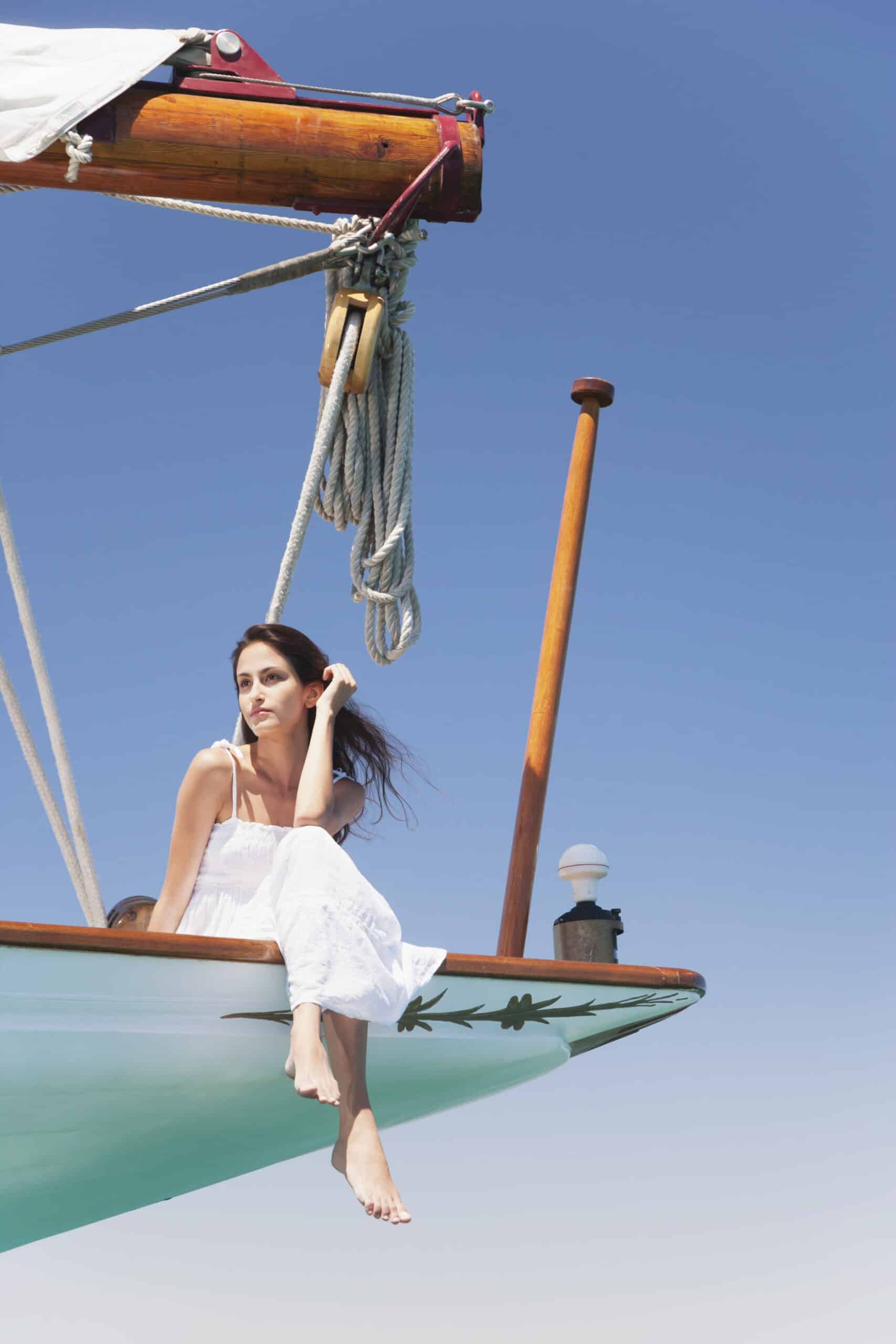 Eine Frau in einem weißen Kleid sitzt auf dem Bug eines Segelboots und lässt ihre Füße über die Kante baumeln. Sie blickt nachdenklich in die Ferne. Der Himmel ist klar und blau, und Teile der Holzkonstruktion und der Seile des Bootes sind sichtbar. © Fotografie Tomas Rodriguez