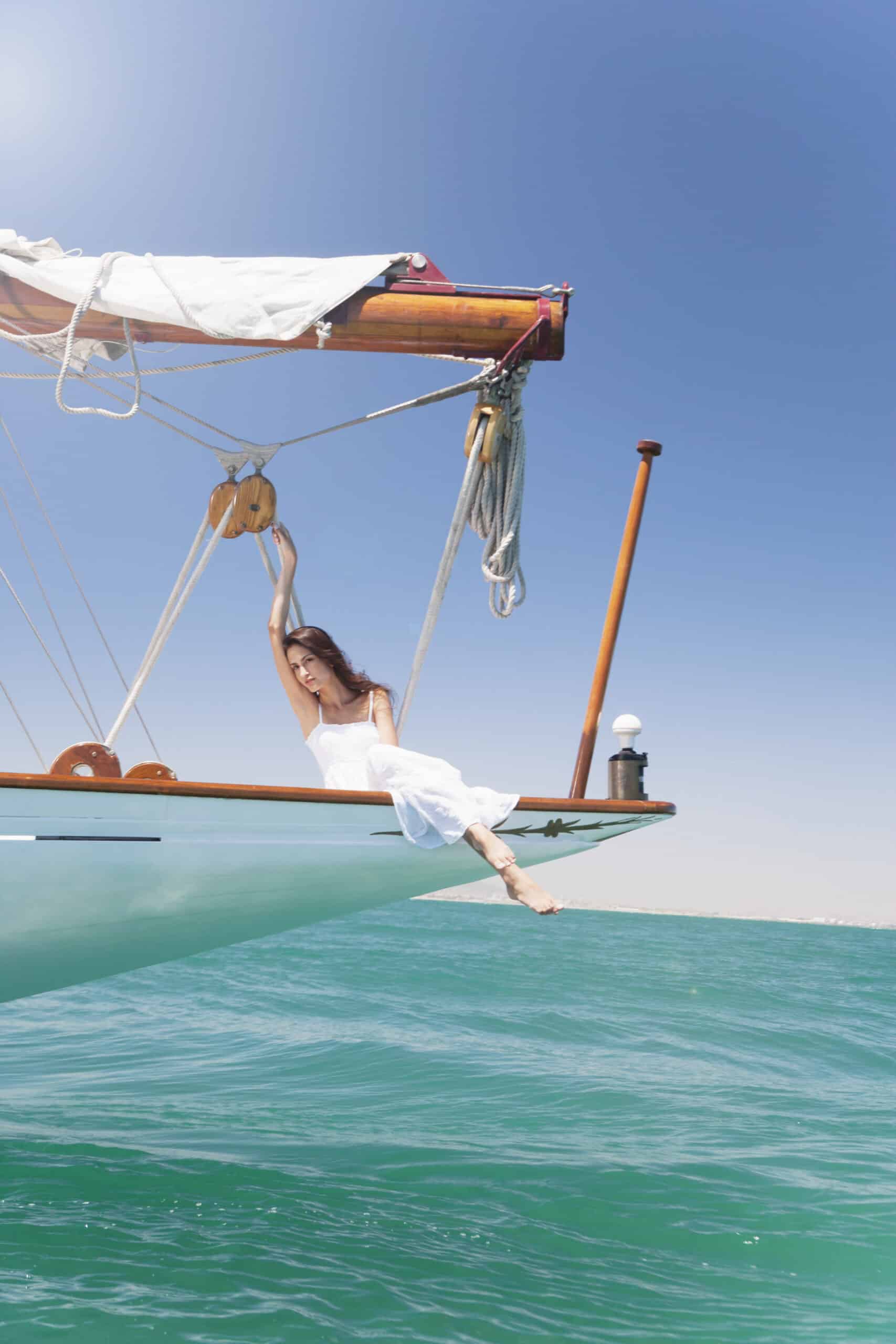 Eine Frau in einem weißen Kleid hängt verspielt am Bugkorb eines im Meer treibenden Segelboots. Der strahlend blaue Himmel und das Meer bilden einen ruhigen, lebendigen Hintergrund. Auch die Holzkonstruktion und die Takelage des Segelboots sind vor dem klaren Himmel gut zu erkennen. © Fotografie Tomas Rodriguez