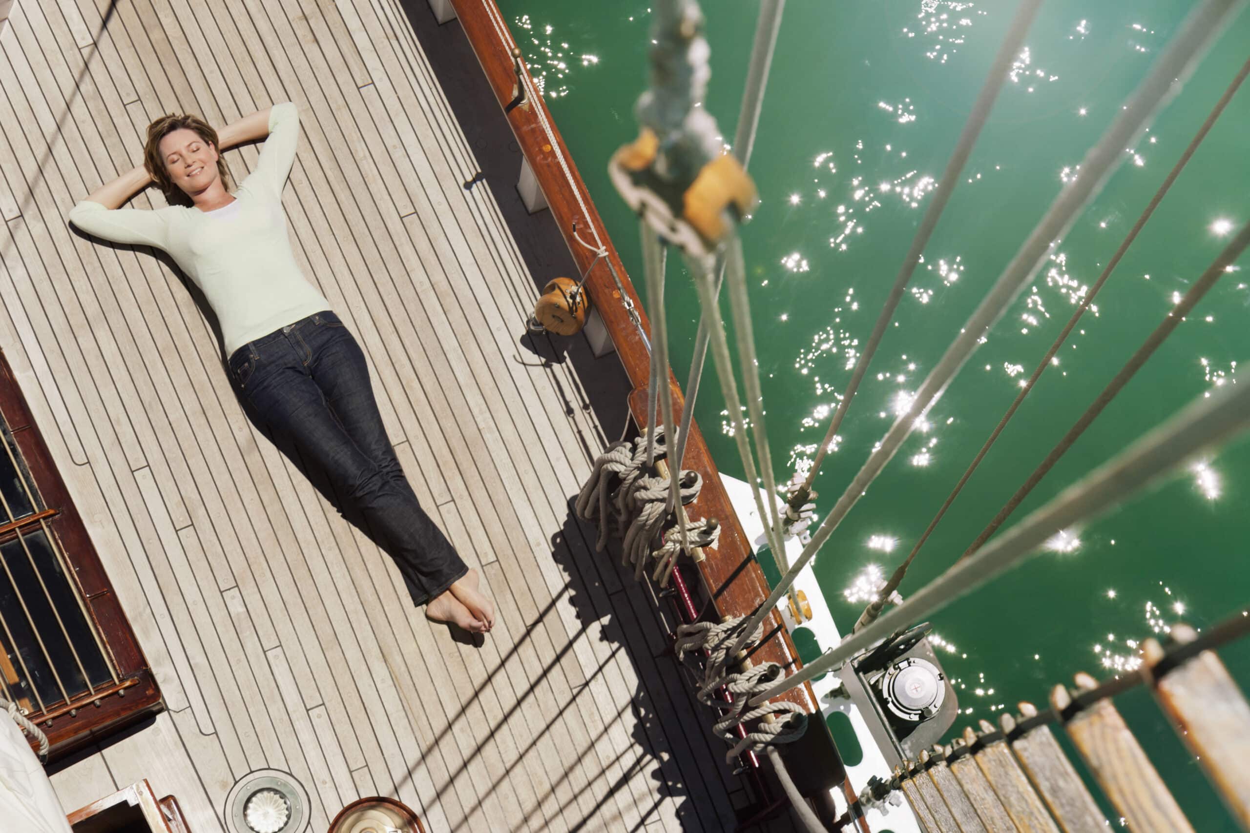 Eine Person liegt auf dem Rücken und entspannt sich mit verschränkten Armen auf dem Holzdeck eines Segelboots. Das Boot liegt im grünen, sonnenbeschienenen Wasser und an der Seite des Decks sind verschiedene Seile und Takelage sichtbar. © Fotografie Tomas Rodriguez