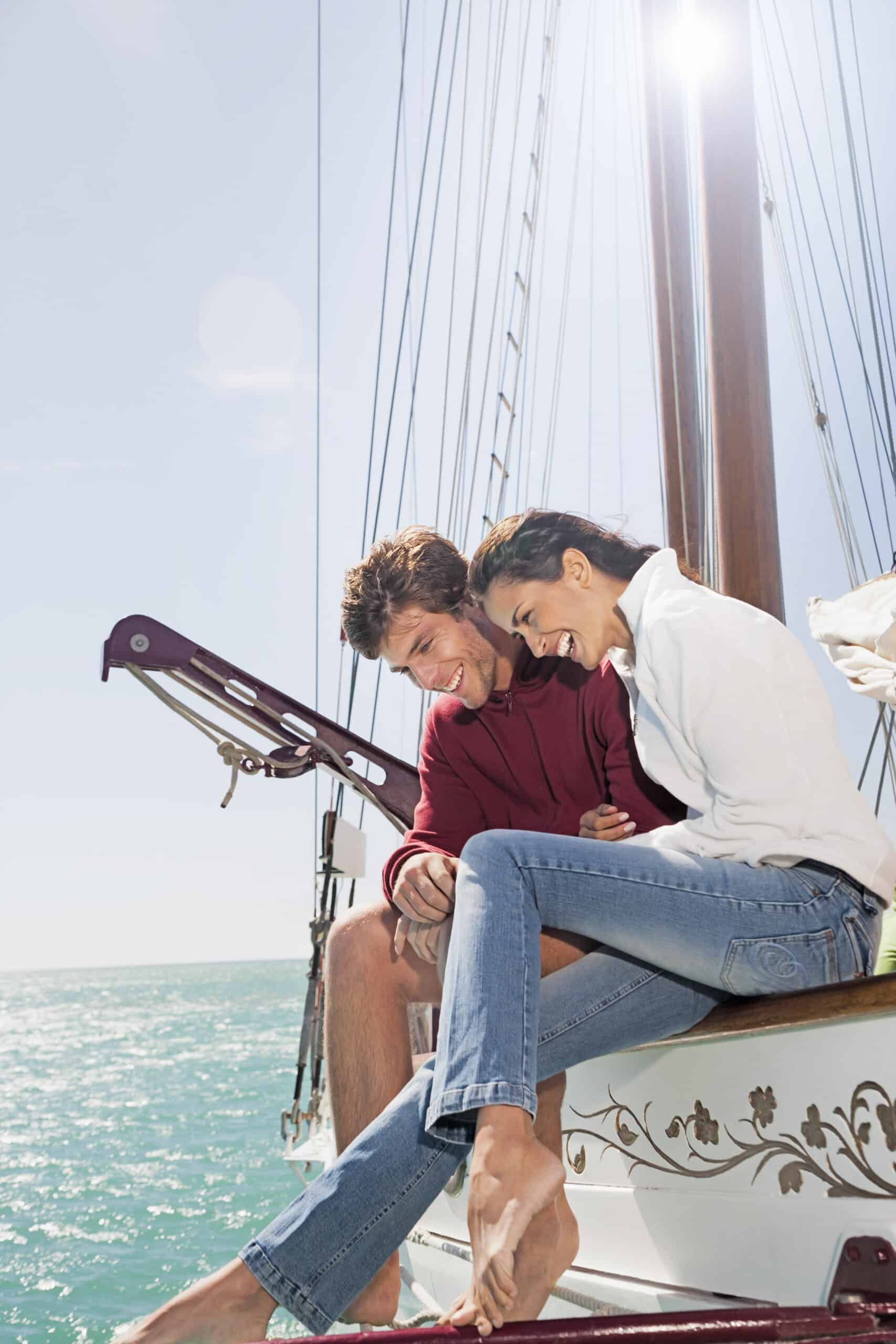 Ein Mann und eine Frau sitzen lächelnd und lachend am Rand eines Segelboots. Sie sind leger gekleidet; der Mann trägt einen roten Kapuzenpullover und die Frau ein weißes Hemd und Jeans. Die Sonne scheint hell und im Hintergrund ist das Meer ruhig und blau. © Fotografie Tomas Rodriguez