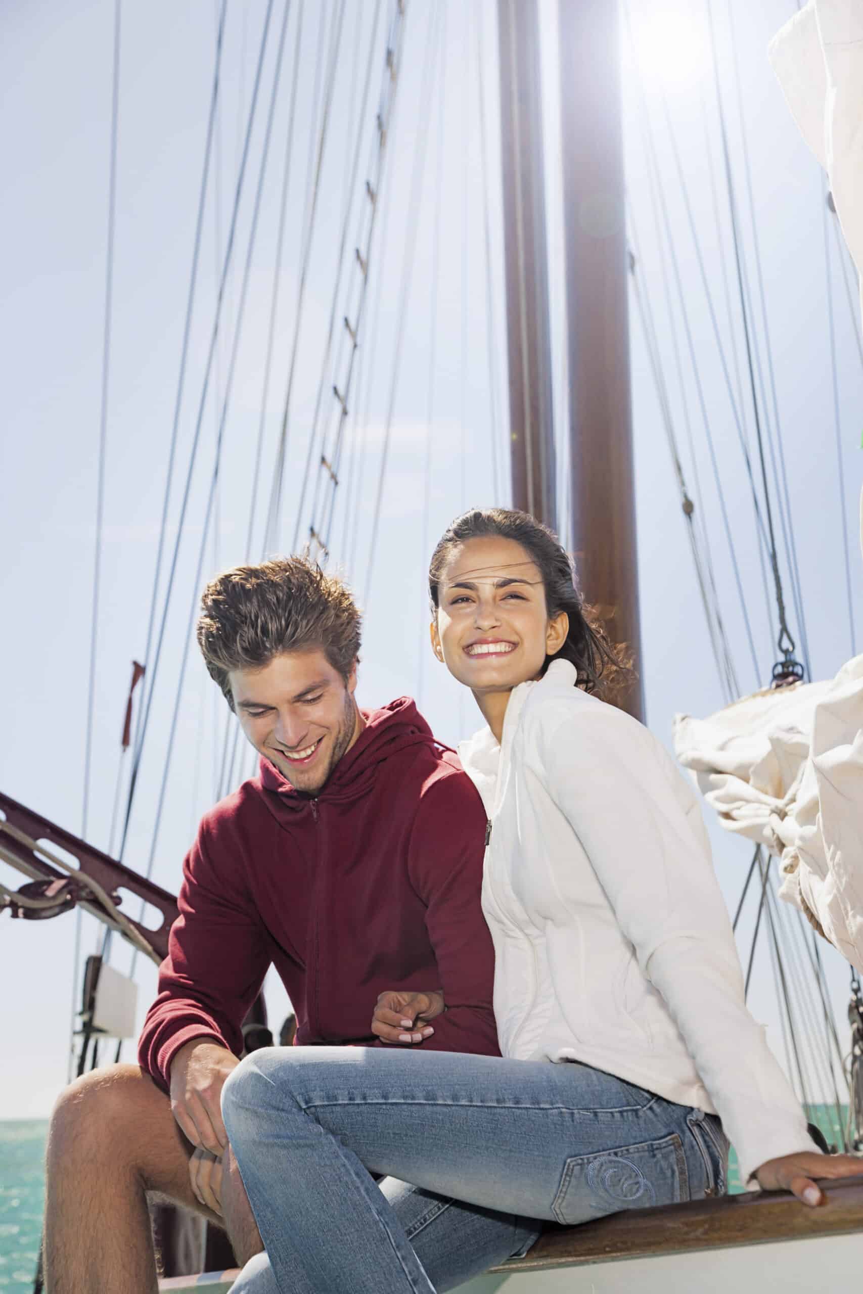 Ein Mann und eine Frau sitzen auf dem Deck eines Segelboots. Der Mann, der einen roten Kapuzenpullover trägt, schaut nach unten und lächelt, während die Frau, die eine weiße Jacke und blaue Jeans trägt, strahlend in die Kamera lächelt. Der Himmel ist klar und sonnig, und der Mast und die Segel des Bootes sind sichtbar. © Fotografie Tomas Rodriguez