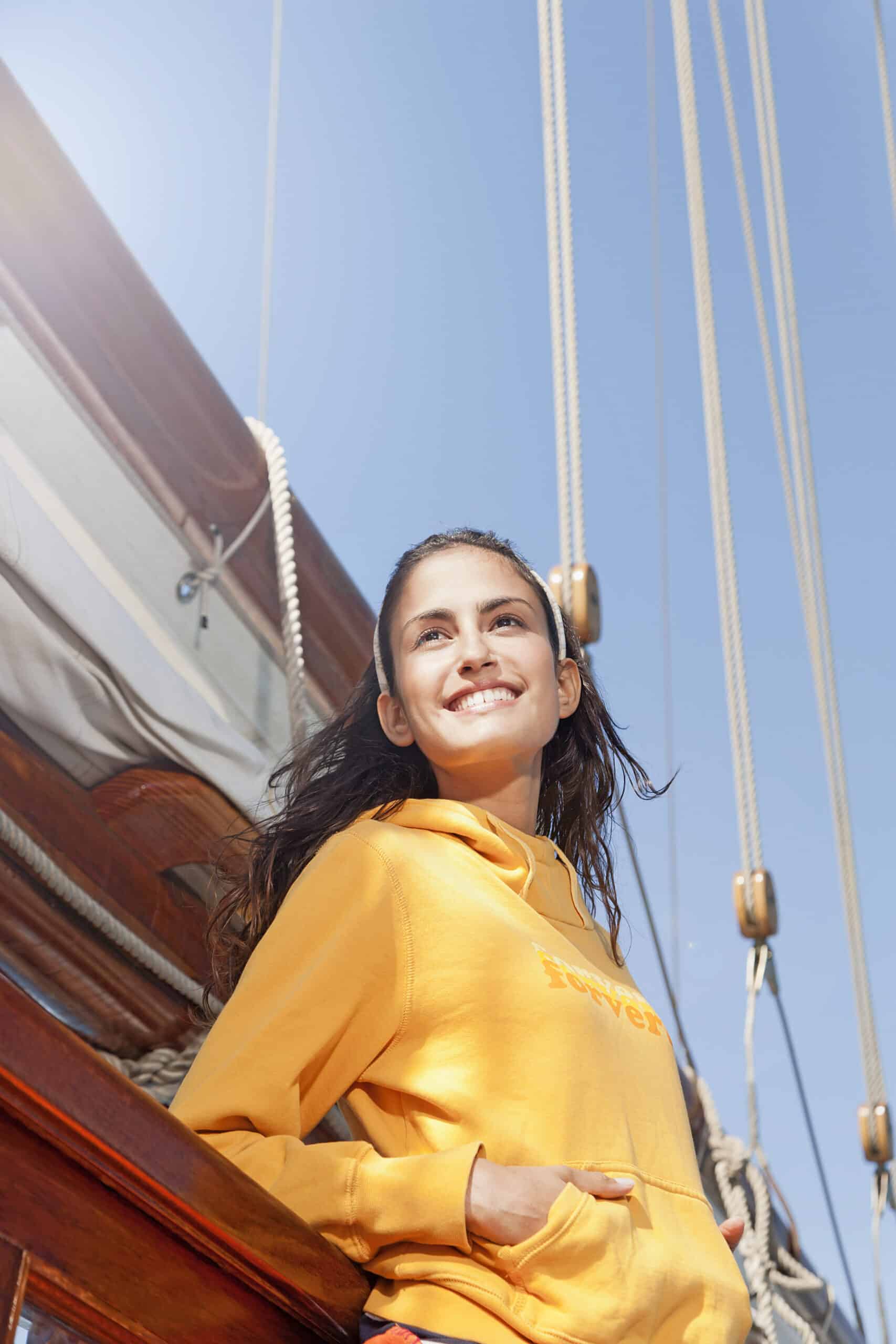 Eine junge Frau in einem gelben Kapuzenpullover steht an einem sonnigen Tag auf dem Deck eines Segelboots. Sie hat ihre Hände in der Kapuzentasche und lächelt und blickt in die Ferne. Hinter ihr sind die Takelage und Seile des Bootes vor einem klaren blauen Himmel zu sehen. © Fotografie Tomas Rodriguez