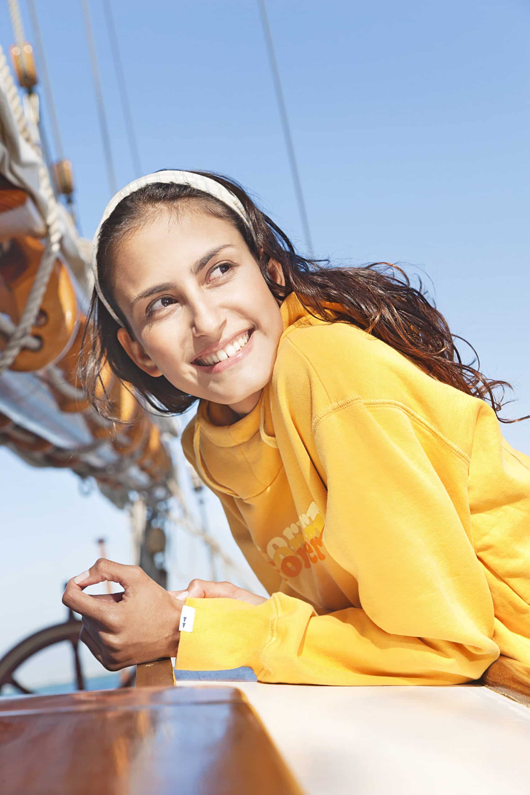 Eine lächelnde Person mit langen braunen Haaren und einem weißen Stirnband lehnt auf dem Deck eines Segelboots. Sie trägt einen leuchtend gelben Kapuzenpullover und blickt in die Ferne. Im Hintergrund sind die Takelage des Bootes und ein klarer blauer Himmel zu sehen. © Fotografie Tomas Rodriguez