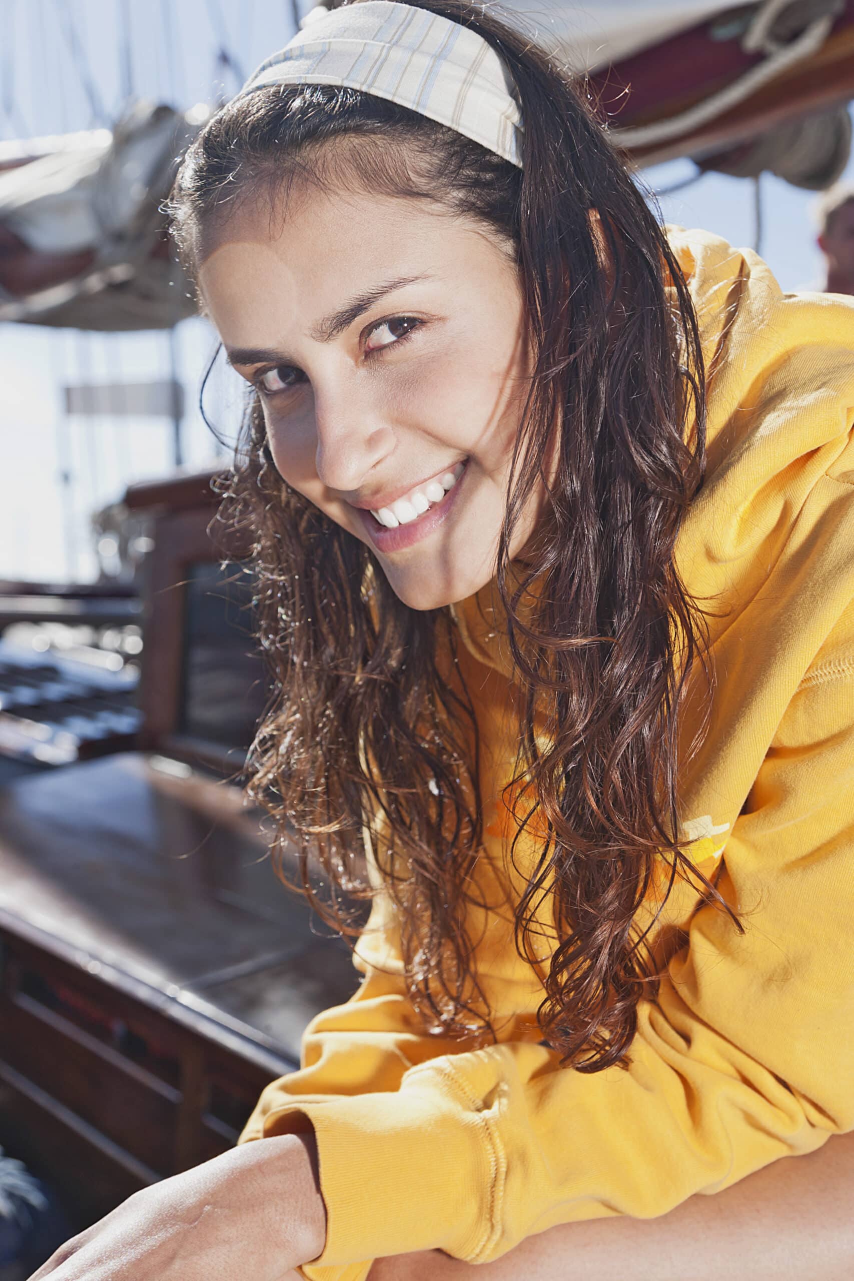 Eine junge Frau mit langen braunen Haaren, einem gelben Kapuzenpullover und einem karierten Stirnband, lächelt in die Kamera. Sie ist auf einem Boot mit nautischer Ausrüstung im Hintergrund und genießt einen sonnigen Tag. © Fotografie Tomas Rodriguez