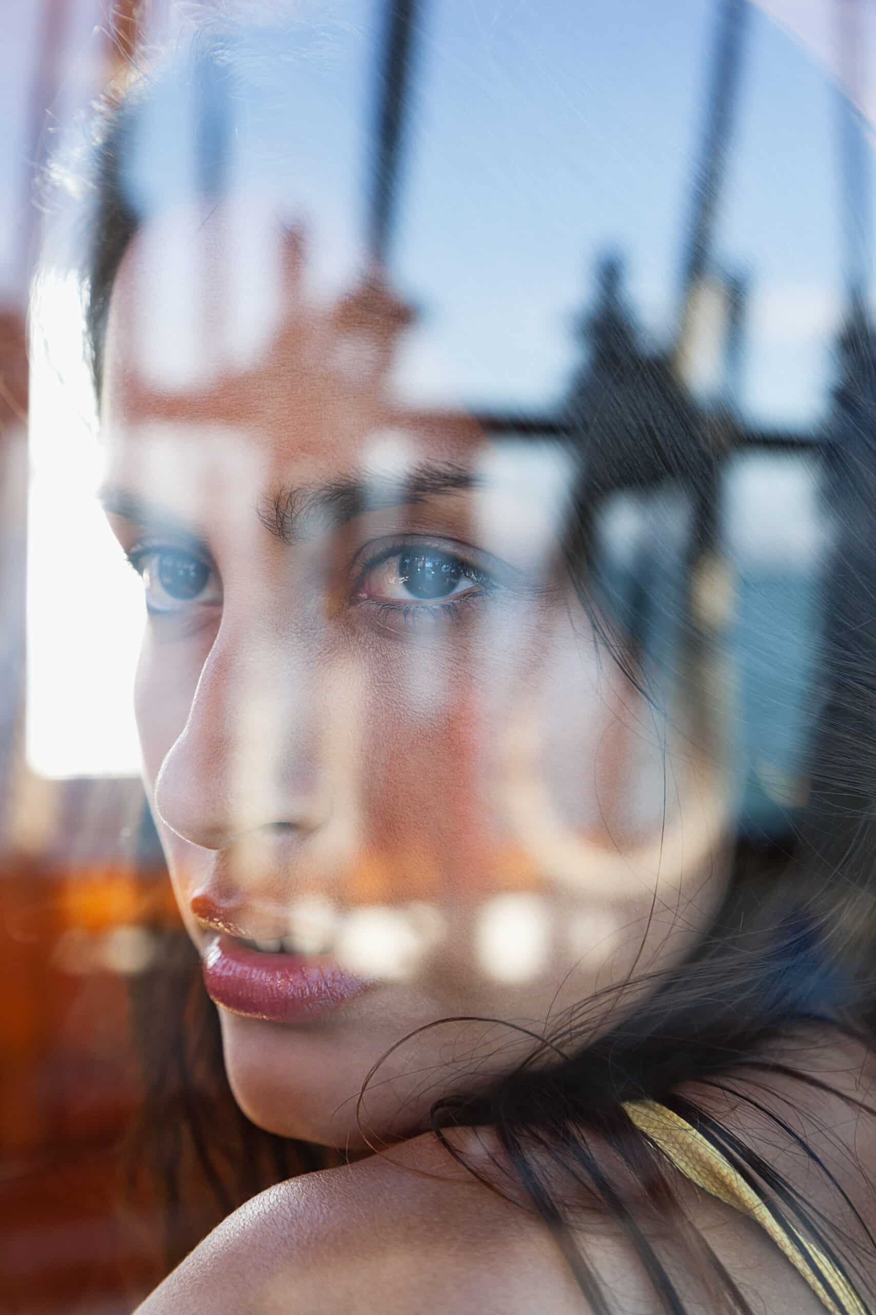 Eine Nahaufnahme einer Frau mit langem dunklem Haar, die über ihre Schulter blickt, gesehen durch ein Fenster mit Spiegelungen eines Gebäudes und des Himmels. Das Bild fängt einen intensiven und nachdenklichen Ausdruck auf ihrem Gesicht ein, hervorgehoben durch natürliches Licht. © Fotografie Tomas Rodriguez