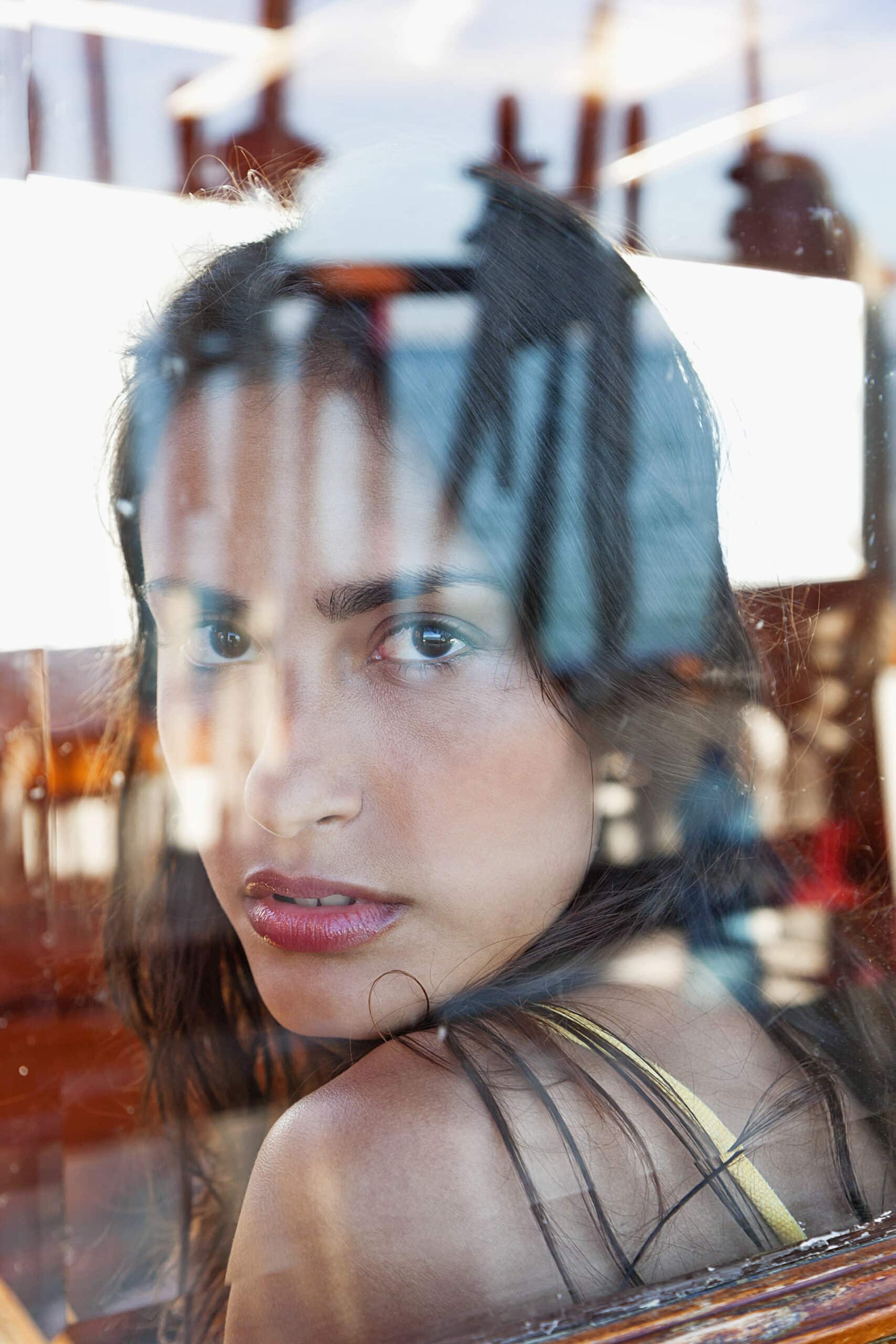 Eine Frau mit langem dunklem Haar und ernstem Gesichtsausdruck blickt durch ein Glasfenster. Auf dem Glas sind Spiegelungen der Fensterscheiben und einer sonnenbeschienenen Außenszene zu sehen, die ihr Gesicht teilweise verdecken. Sie trägt ein gelbes Oberteil und hat einen nachdenklichen Blick. © Fotografie Tomas Rodriguez