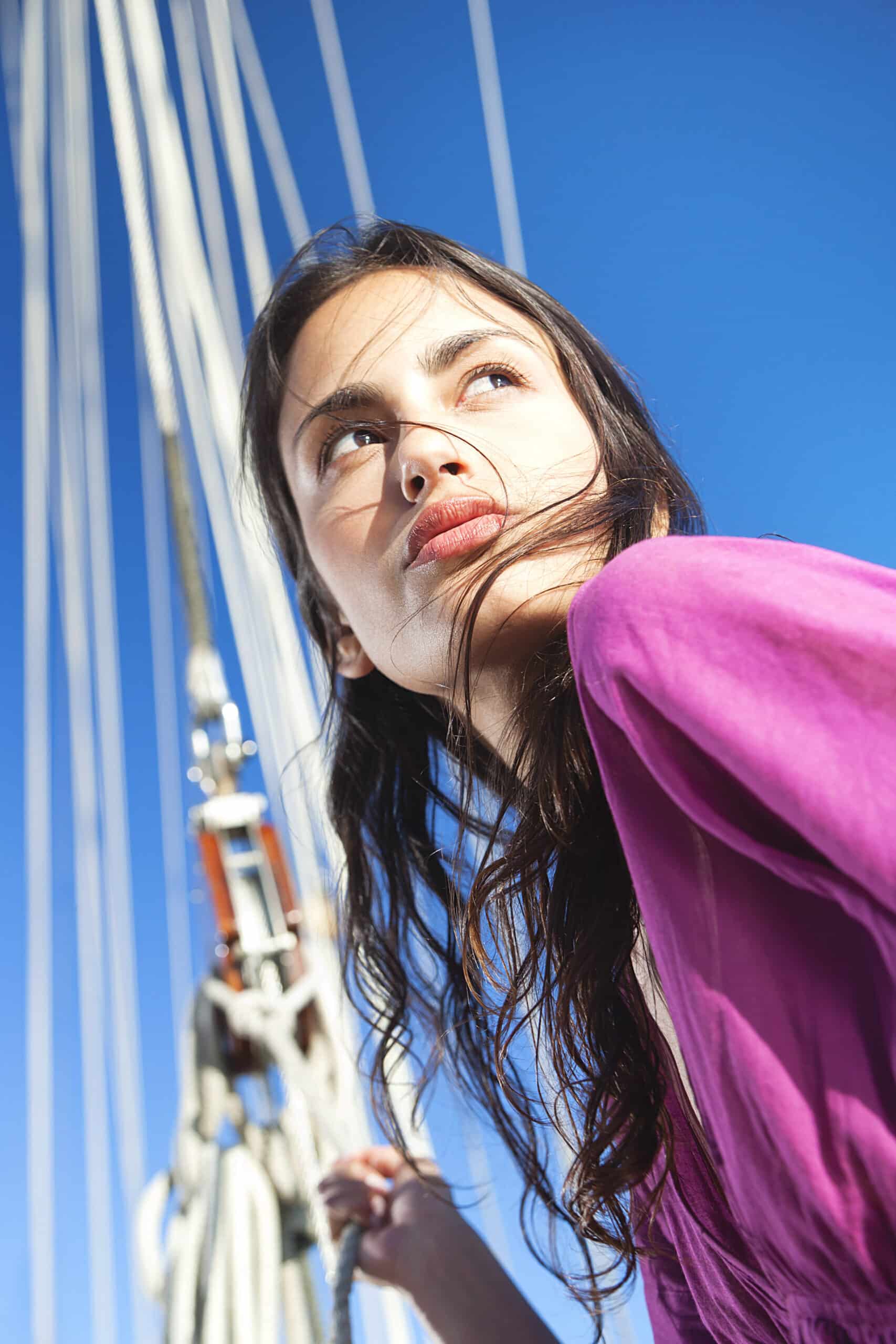 Eine Person mit langem, dunklem Haar und einem lila Oberteil blickt an einem sonnigen Tag in die Ferne. Sie steht neben einigen Segeln und Takelage vor einem klaren blauen Himmel. © Fotografie Tomas Rodriguez