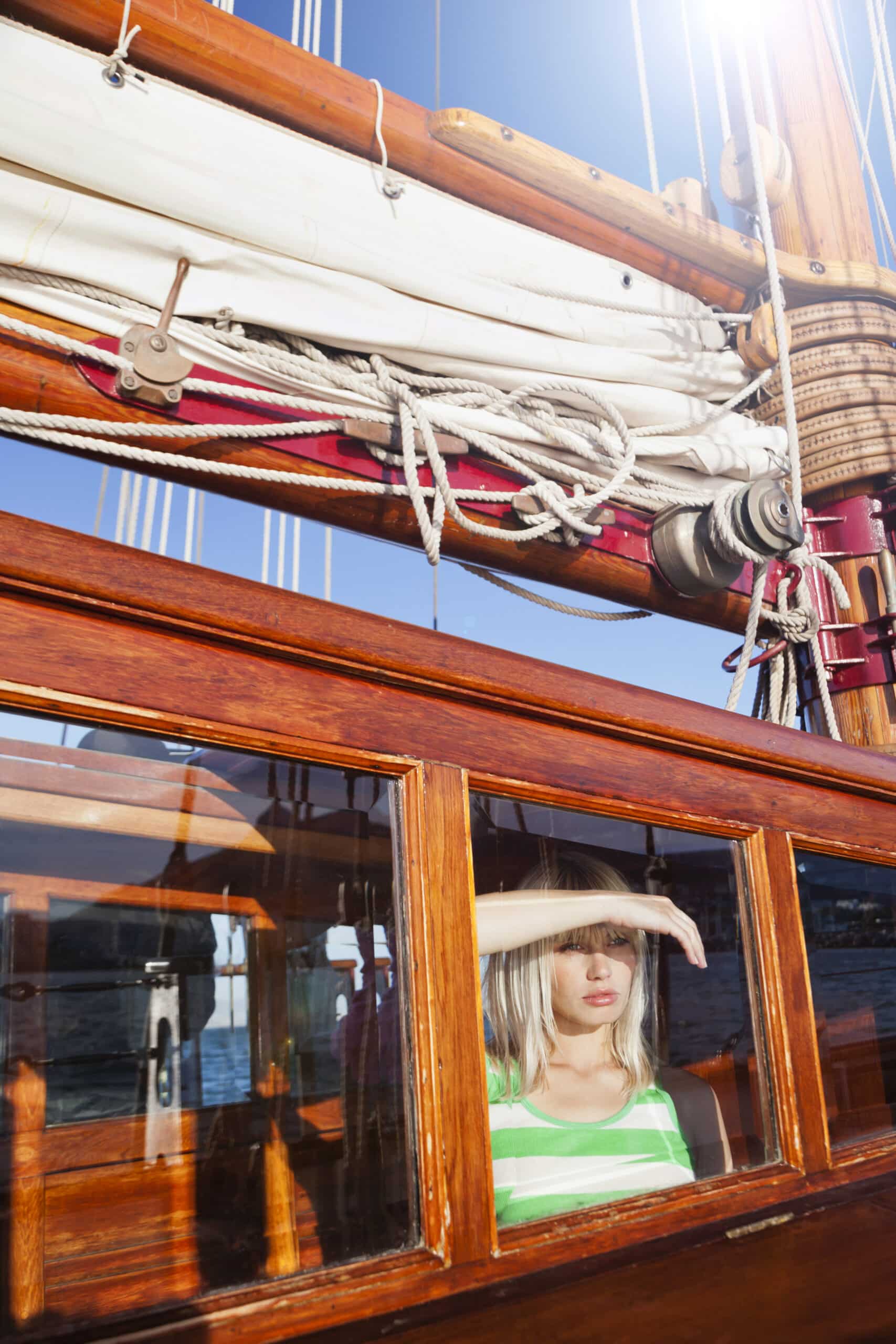 Eine Person mit blondem Haar und einem grün-weiß gestreiften Tanktop steht auf einem Segelboot und wird durch eine erhobene Hand vor der Sonne geschützt. Sie ist teilweise durch das hölzerne Kabinenfenster des Bootes zu sehen, über ihnen hängen Seile und Segel. © Fotografie Tomas Rodriguez