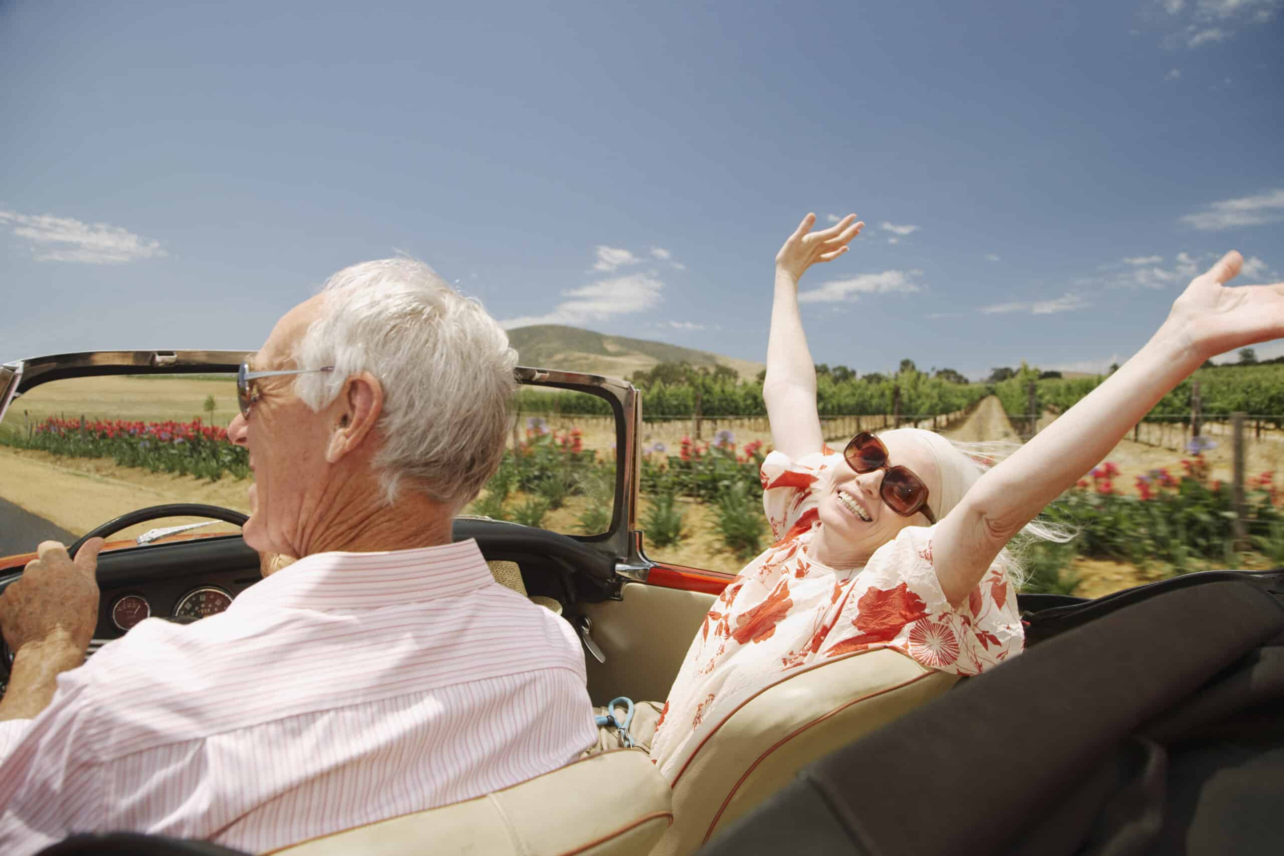 Ein älterer Mann und eine ältere Frau fahren an einem sonnigen Tag in einem roten Cabrio mit offenem Verdeck. Der Mann fährt, während die Frau, die eine Sonnenbrille und ein Kopftuch trägt, lächelt und ihre Arme freudig in die Höhe hebt. Hinter ihnen sind grüne Felder und ein klarer blauer Himmel. © Fotografie Tomas Rodriguez