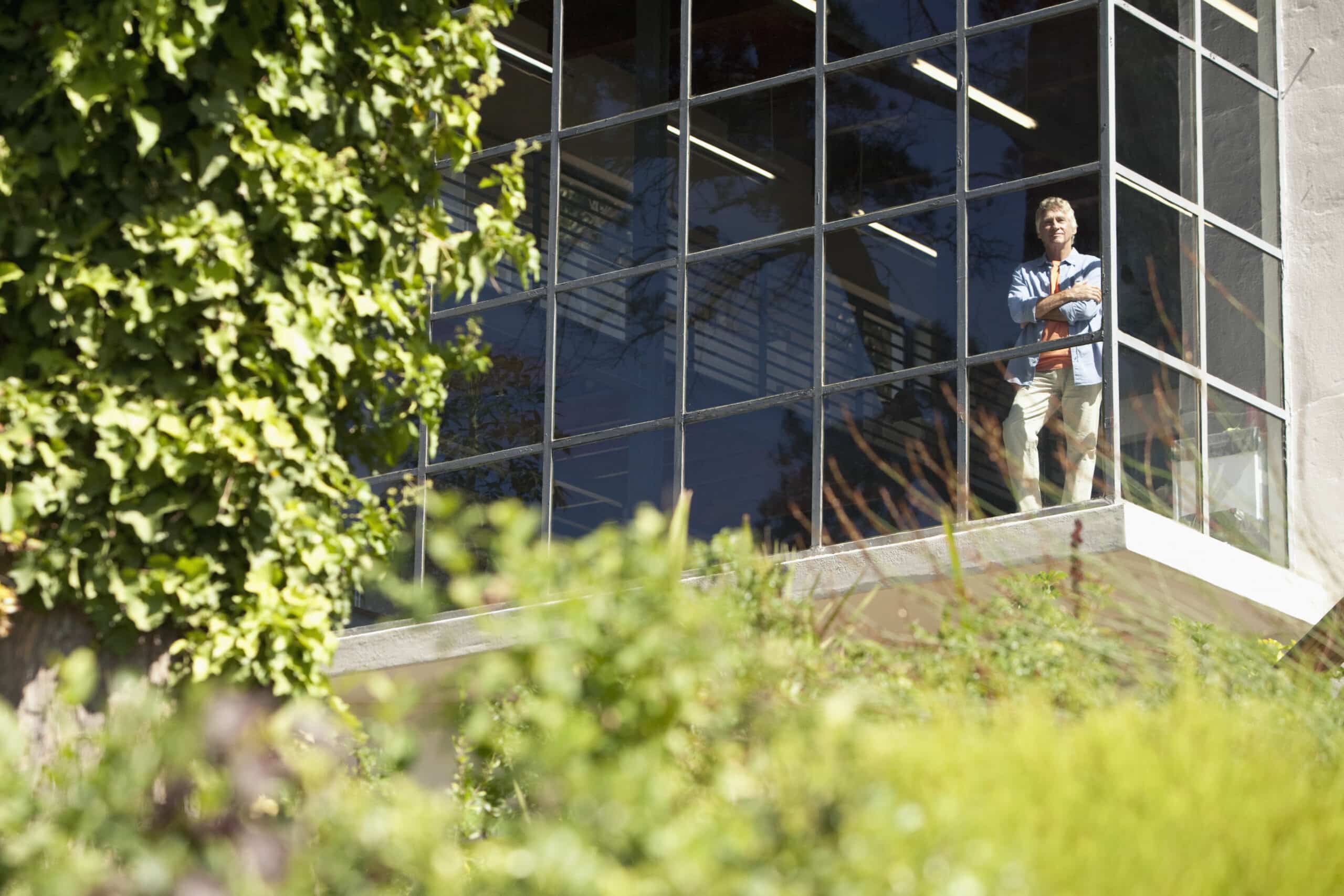Eine Person steht in einem Raum mit großen, deckenhohen Fenstern und blickt nach draußen. Der Blick durch die Fenster umfasst das Grün der Bäume und Pflanzen draußen. Die Person wirkt nachdenklich, hat verschränkte Arme und trägt Freizeitkleidung. © Fotografie Tomas Rodriguez