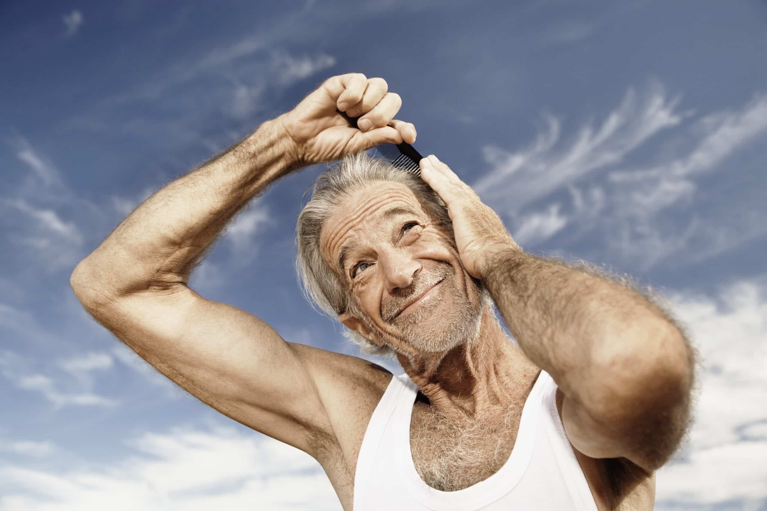 Ein älterer, muskulöser Mann in einem weißen Tanktop steht draußen unter einem klaren blauen Himmel. Er lächelt und kämmt mit seiner rechten Hand sein weißes Haar mit einem schwarzen Kamm. Er sieht fröhlich und zufrieden aus. © Fotografie Tomas Rodriguez