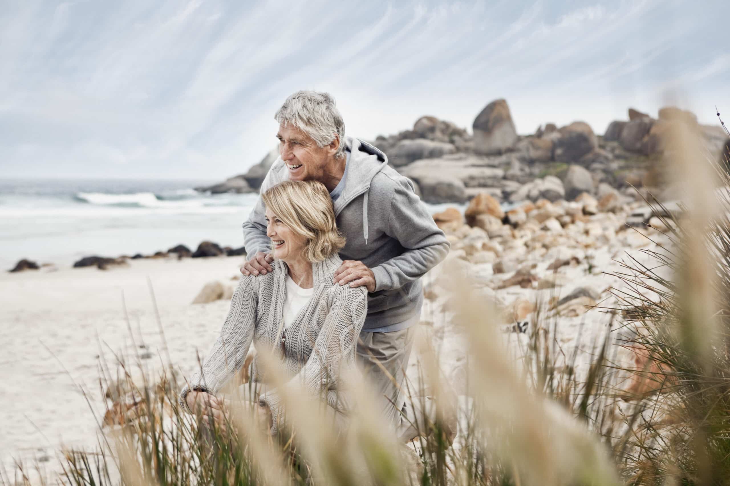 Ein älteres Paar genießt die gemeinsame Zeit am Strand. Beide lächeln und tragen legere, warme Kleidung. Im Vordergrund des Strandes sind Felsen und Gras zu sehen, im Hintergrund ein ruhiges Meer und ein bewölkter Himmel. © Fotografie Tomas Rodriguez