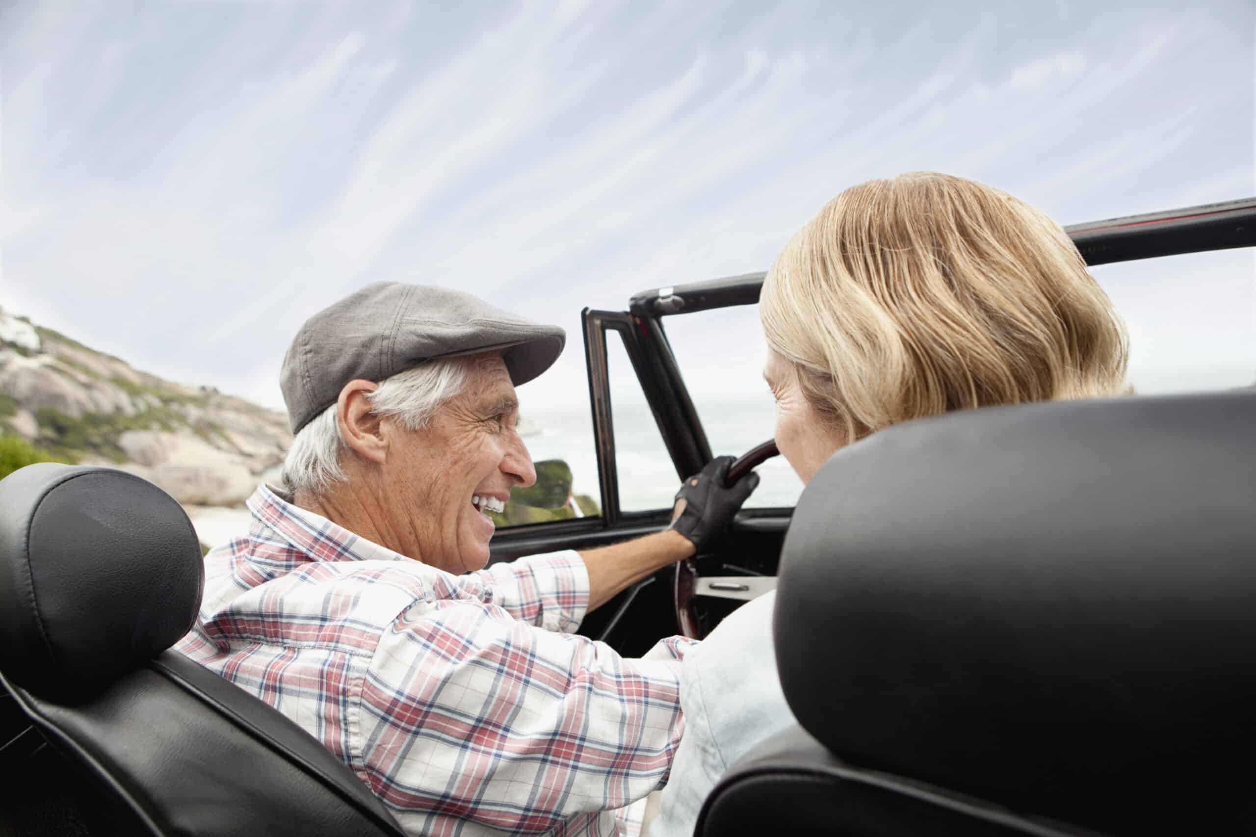 Ein älterer Mann und eine ältere Frau genießen eine Fahrt in einem Cabrio mit offenem Verdeck. Der Mann, der ein kariertes Hemd und eine Schiebermütze trägt, lächelt die Frau an, die blondes Haar hat. Im Hintergrund sind ein klarer, blauer Himmel und eine verschwommene Szenerie zu sehen, die auf Bewegung hindeutet. © Fotografie Tomas Rodriguez