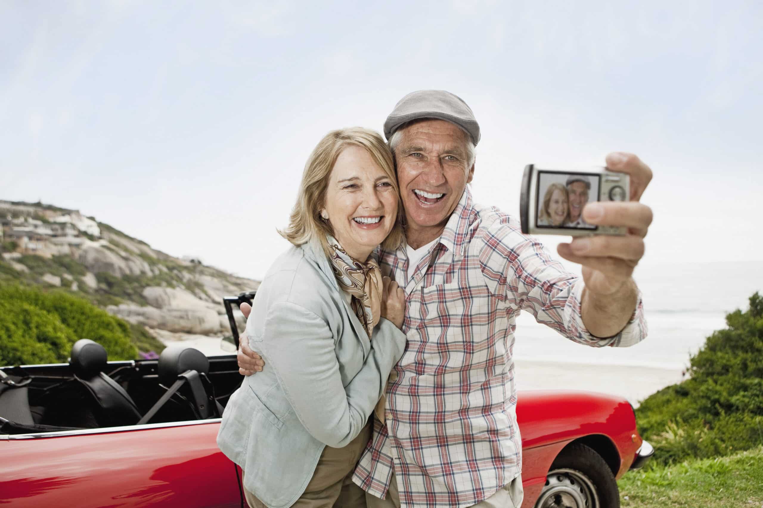 Ein älteres Paar steht neben einem roten Cabrio und hat einen malerischen Meerblick. Der Mann, der ein kariertes Hemd und eine Mütze trägt, hält eine Kamera für ein Selfie. Beide lächeln herzlich, und die Frau in einer hellen Jacke umarmt den Mann. Ein heller und sonniger Tag. © Fotografie Tomas Rodriguez