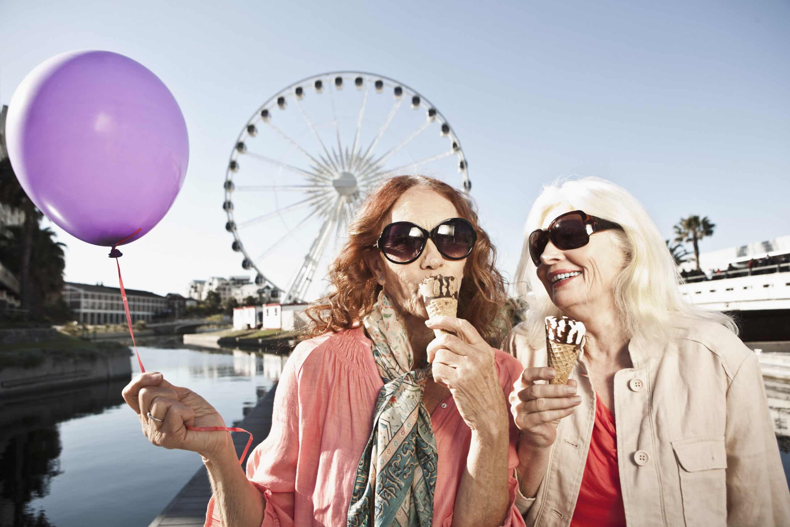 Zwei lächelnde ältere Frauen mit Sonnenbrillen essen an einem sonnigen Tag Eis. Eine hält einen lila Luftballon. Im Hintergrund sind ein großes Riesenrad und ein klarer blauer Himmel zu sehen, die sich im ruhigen Wasser eines nahe gelegenen Kanals spiegeln. Sie wirken glücklich und entspannt. © Fotografie Tomas Rodriguez