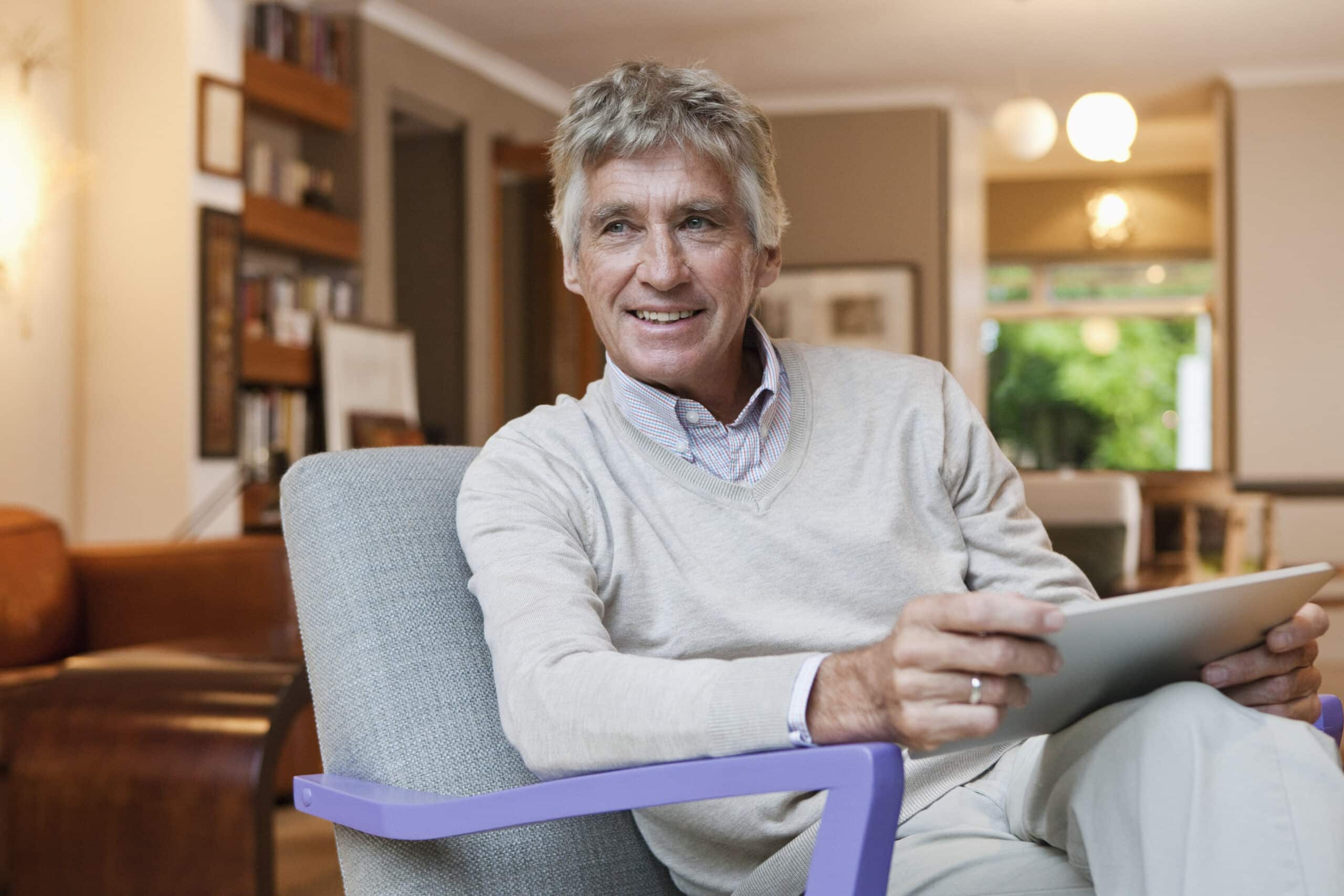 Ein älterer Mann mit grauem Haar, der einen leichten Pullover über einem Hemd mit Kragen trägt, sitzt in einem modernen Wohnzimmer. Er lächelt sanft und hält ein Tablet in den Händen. Der Raum ist warm beleuchtet und verfügt über eine Mischung aus zeitgenössischen und klassischen Dekorelementen, darunter Bücherregale. © Fotografie Tomas Rodriguez