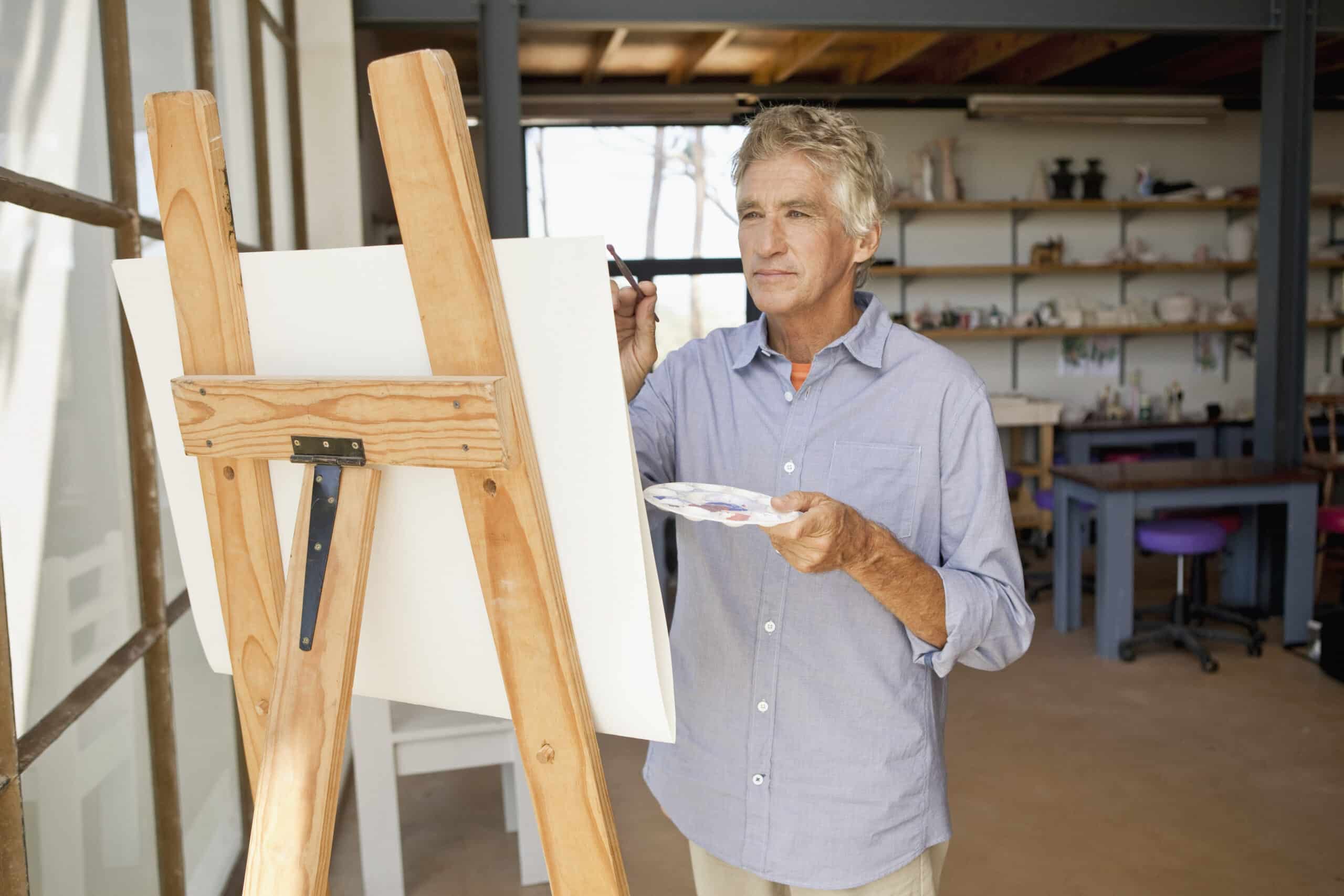 Ein älterer Mann mit grauem Haar malt an einer Staffelei in einem geräumigen, gut beleuchteten Kunstatelier auf einer Leinwand. In der einen Hand hält er eine Palette mit verschiedenen Farben und in der anderen einen Pinsel und konzentriert sich auf sein Kunstwerk. Im Hintergrund sind Regale und Tische zu sehen. © Fotografie Tomas Rodriguez