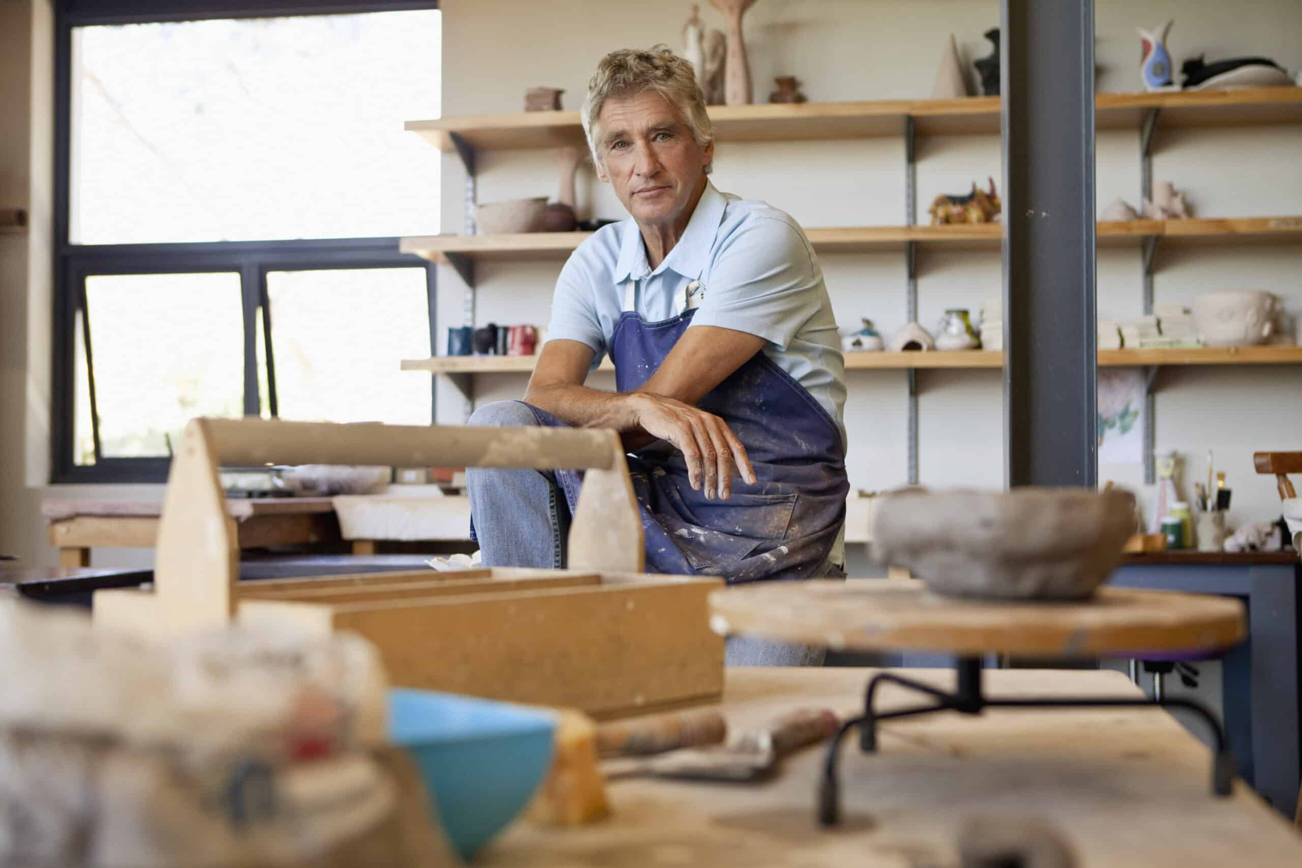 Eine Person mit einer blauen Schürze sitzt auf einem Hocker in einem Töpferstudio. Sie hat einen konzentrierten Gesichtsausdruck und ist von Töpferwerkzeugen und Keramik umgeben. Im Hintergrund sind Regale mit verschiedenen Töpferwaren zu sehen. © Fotografie Tomas Rodriguez