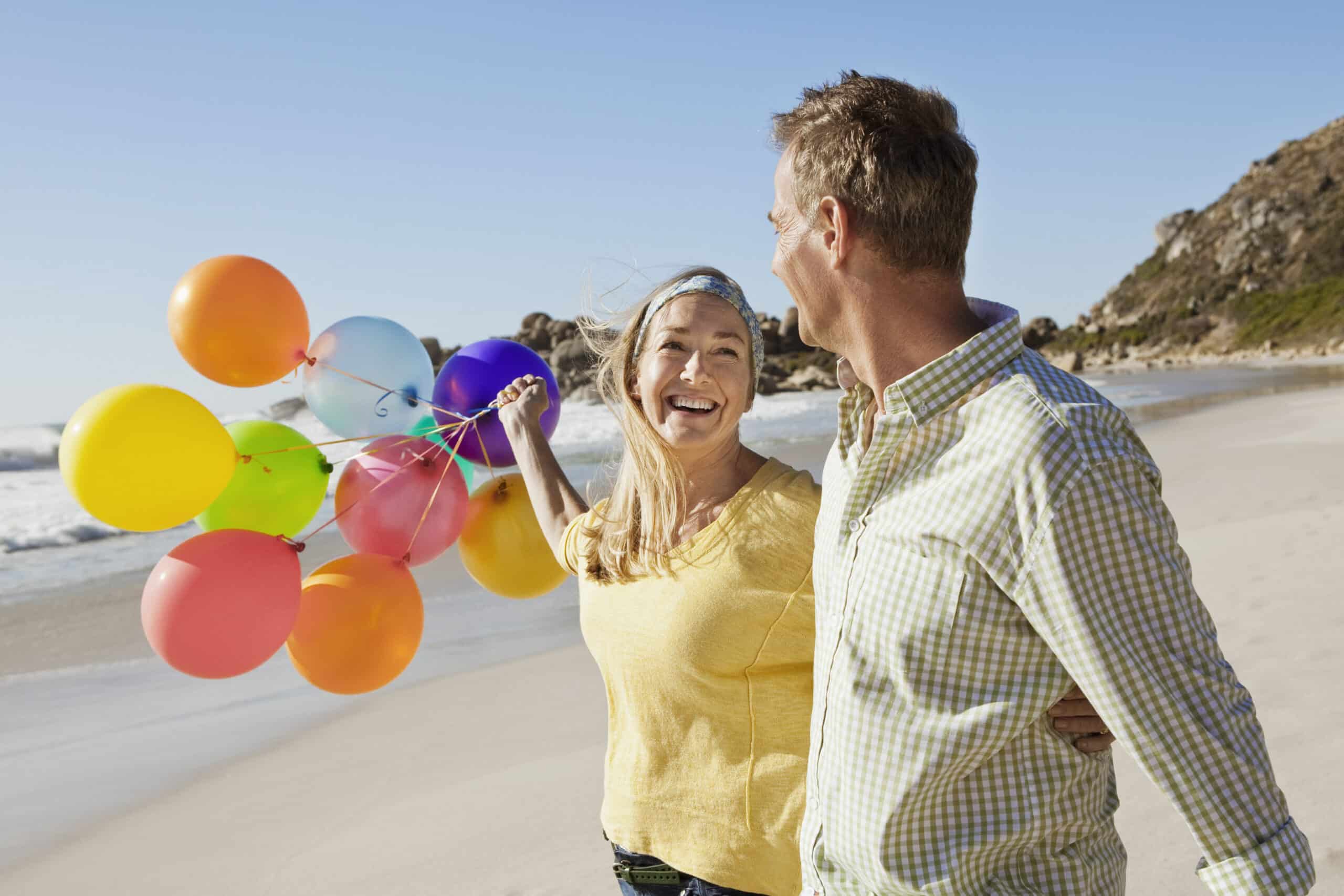 Ein lächelndes Paar geht Arm in Arm an einem sonnigen Strand entlang. Die Frau hält einen Haufen bunter Luftballons. Sie sind leger gekleidet und sehen glücklich aus, mit den Meereswellen und einer felsigen Küste im Hintergrund. © Fotografie Tomas Rodriguez