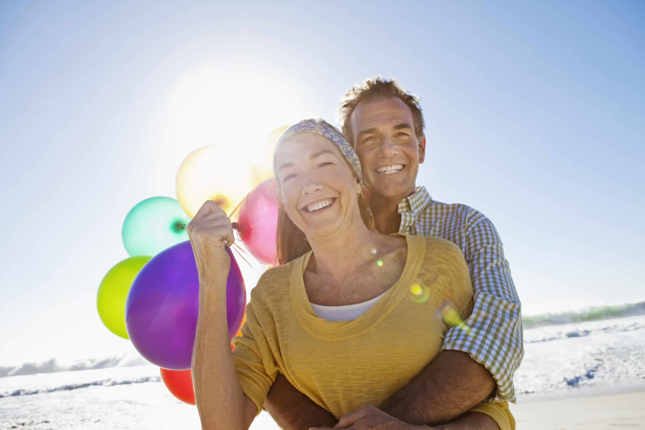 Eine lächelnde ältere Frau in einem gelben Oberteil und einem Stirnband hält bunte Luftballons an einem Strand, während ein Mann hinter ihr sie von hinten umarmt. Die Sonne scheint hell, erzeugt Lichtreflexe und wirft einen warmen Schein auf die Szene. © Fotografie Tomas Rodriguez