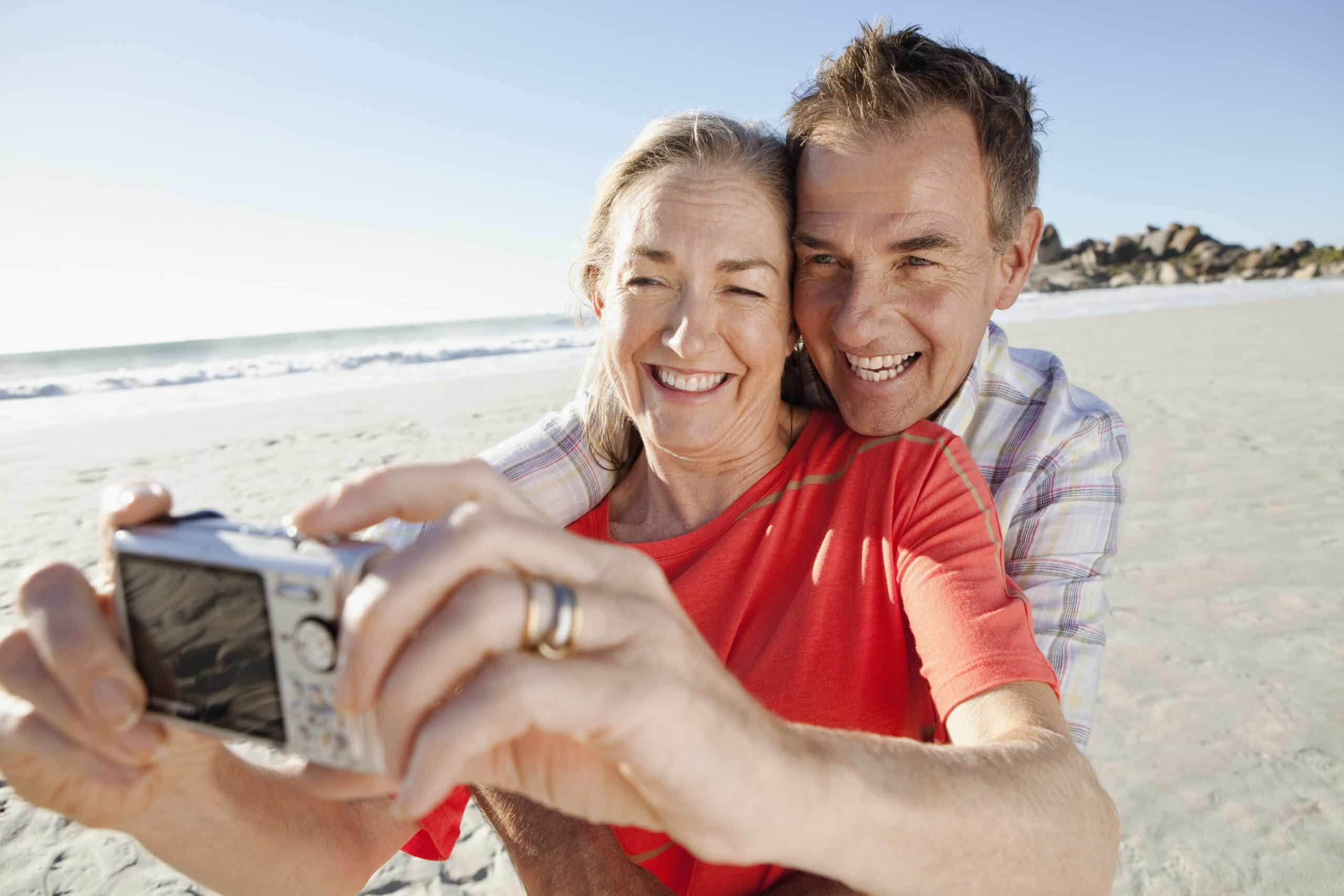 Ein lächelndes älteres Paar umarmt sich, während sie an einem sonnigen Strand mit einer Digitalkamera ein Selfie machen. Die Frau trägt ein rotes Hemd und der Mann ein kariertes Hemd. Der Hintergrund zeigt sandige Küstenlinien und ruhige Meereswellen unter einem klaren blauen Himmel. © Fotografie Tomas Rodriguez