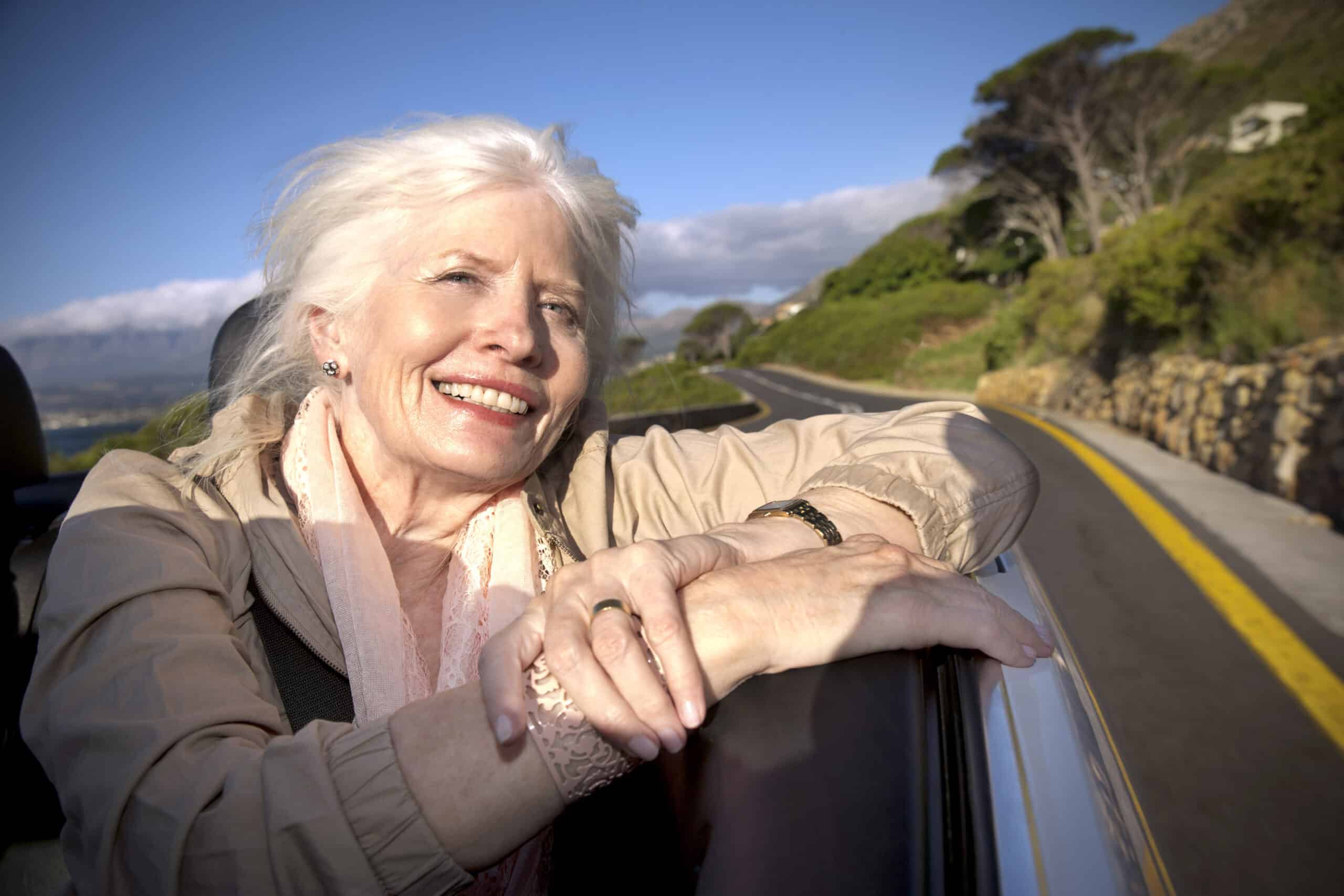 Eine lächelnde ältere Frau mit weißem Haar lehnt sich aus dem Autofenster und stützt ihre Arme auf die Tür. Sie trägt eine helle Jacke und einen Schal. Das Auto fährt auf einer Panoramastraße, die von Grünflächen und Steinmauern gesäumt ist, unter einem strahlend blauen Himmel. © Fotografie Tomas Rodriguez