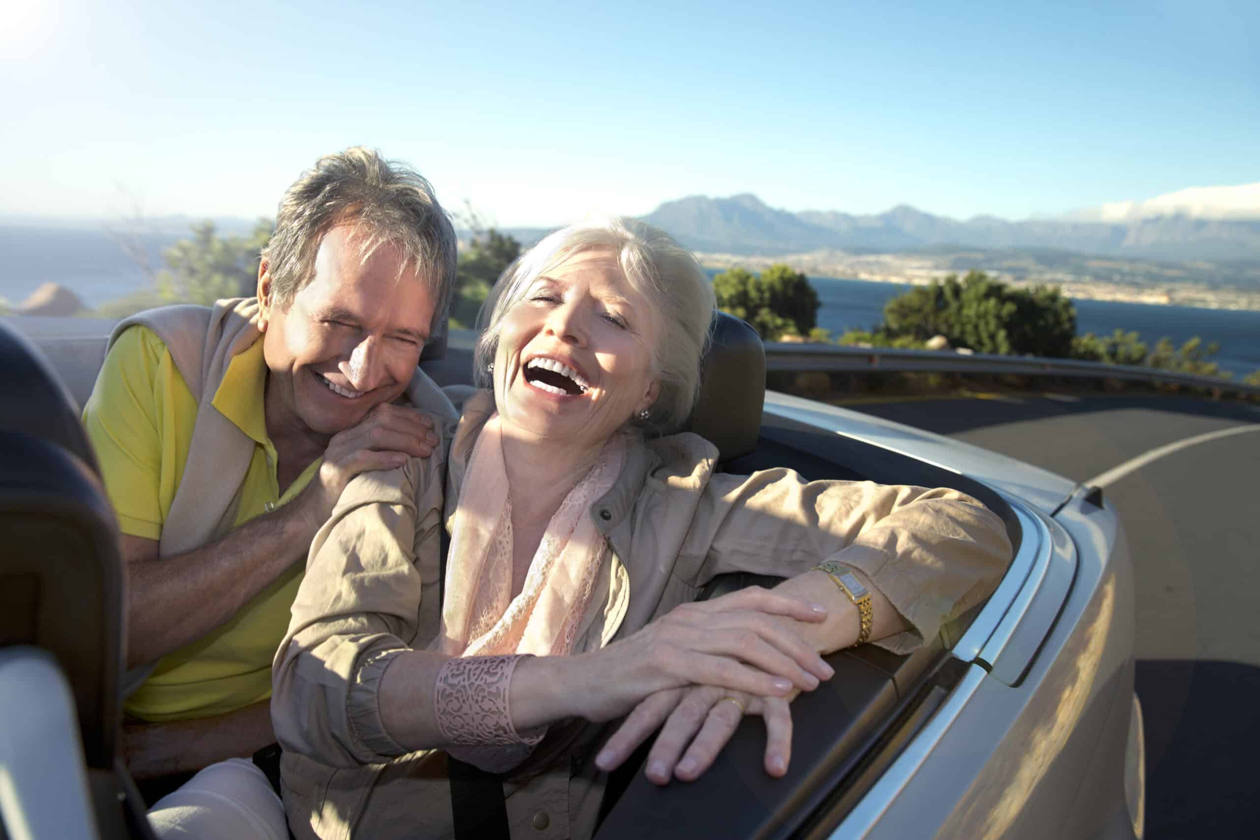 Ein älteres Ehepaar genießt einen freudigen Moment, während es in einem Cabrio auf einer malerischen Küstenstraße sitzt. Beide lächeln und lachen, mit dem Meer, den Bergen und dem klaren blauen Himmel an einem sonnigen Tag im Hintergrund. © Fotografie Tomas Rodriguez