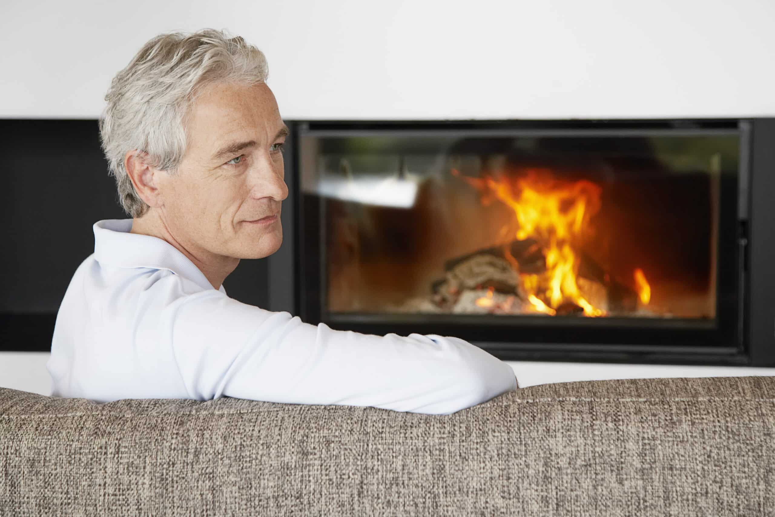 Ein Mann mit kurzen grauen Haaren sitzt auf einem braunen Sofa, leicht nach rechts gedreht, und blickt nachdenklich auf einen brennenden Kamin. Im Inneren des modernen Kamins vor einer weißen Wand sind das Brennholz und die Flammen zu sehen. © Fotografie Tomas Rodriguez