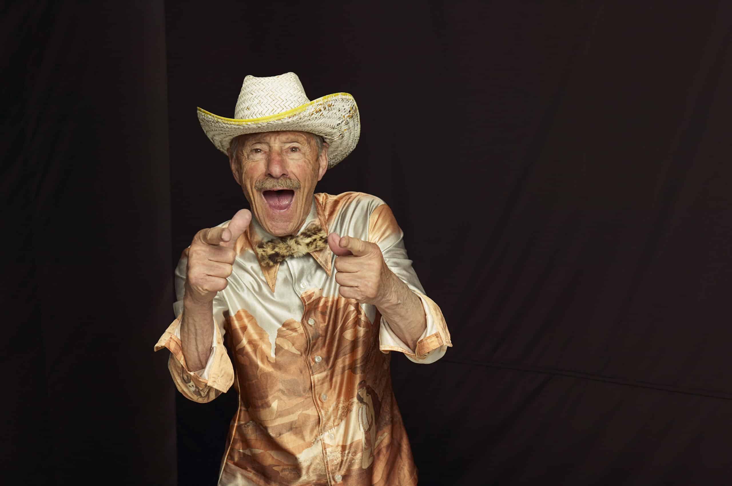 Ein älterer Mann mit einem Cowboyhut aus Stroh, einer beigen Fliege und einem gemusterten Hemd grinst breit und richtet beide Zeigefinger in Richtung Kamera. Der Hintergrund ist dunkel, was den fröhlichen und energiegeladenen Gesichtsausdruck des Mannes noch verstärkt. © Fotografie Tomas Rodriguez