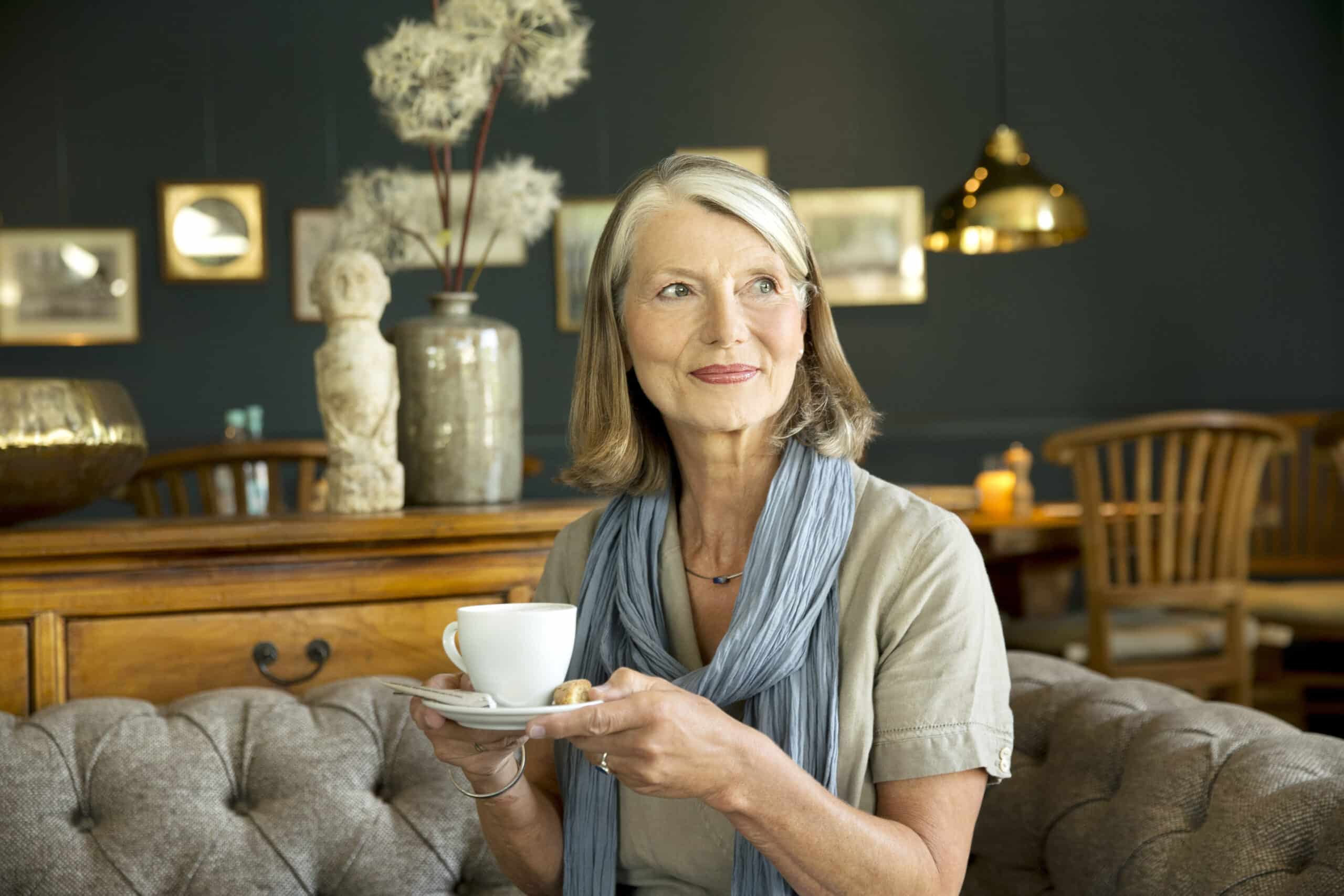 Eine Frau mit schulterlangem grauem Haar, die eine helle Bluse und einen blauen Schal trägt, hält eine weiße Teetasse und Untertasse in der Hand und sitzt auf einem gepolsterten Sofa in einem gemütlichen, warm beleuchteten Raum mit Holzmöbeln und Dekorationsgegenständen. © Fotografie Tomas Rodriguez