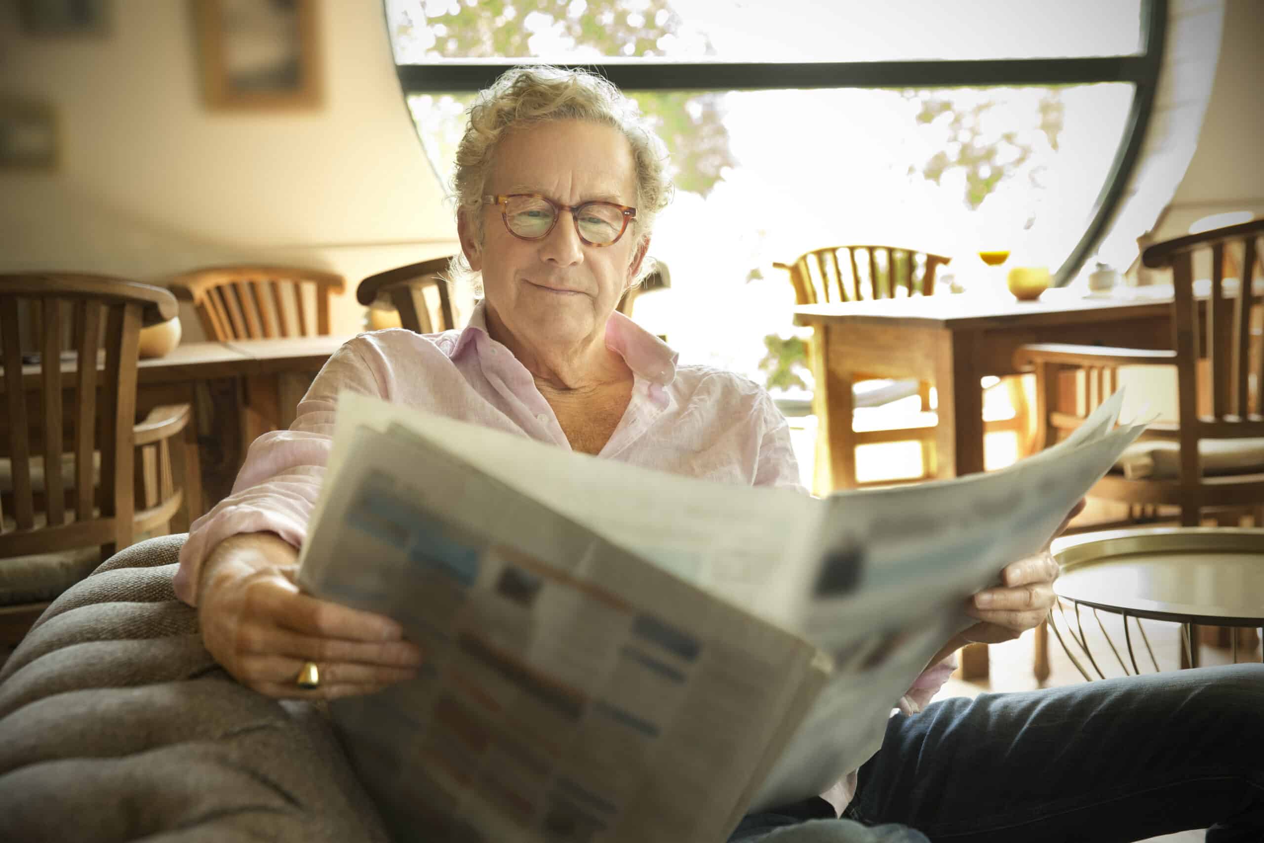 Ein älterer Mann mit Brille sitzt gemütlich auf einem Sofa und liest eine Zeitung. Er trägt ein hellrosa Hemd und im Hintergrund ist ein gemütlicher Raum mit Holzmöbeln und einem großen runden Fenster zu sehen, durch das natürliches Licht hereinfällt. © Fotografie Tomas Rodriguez