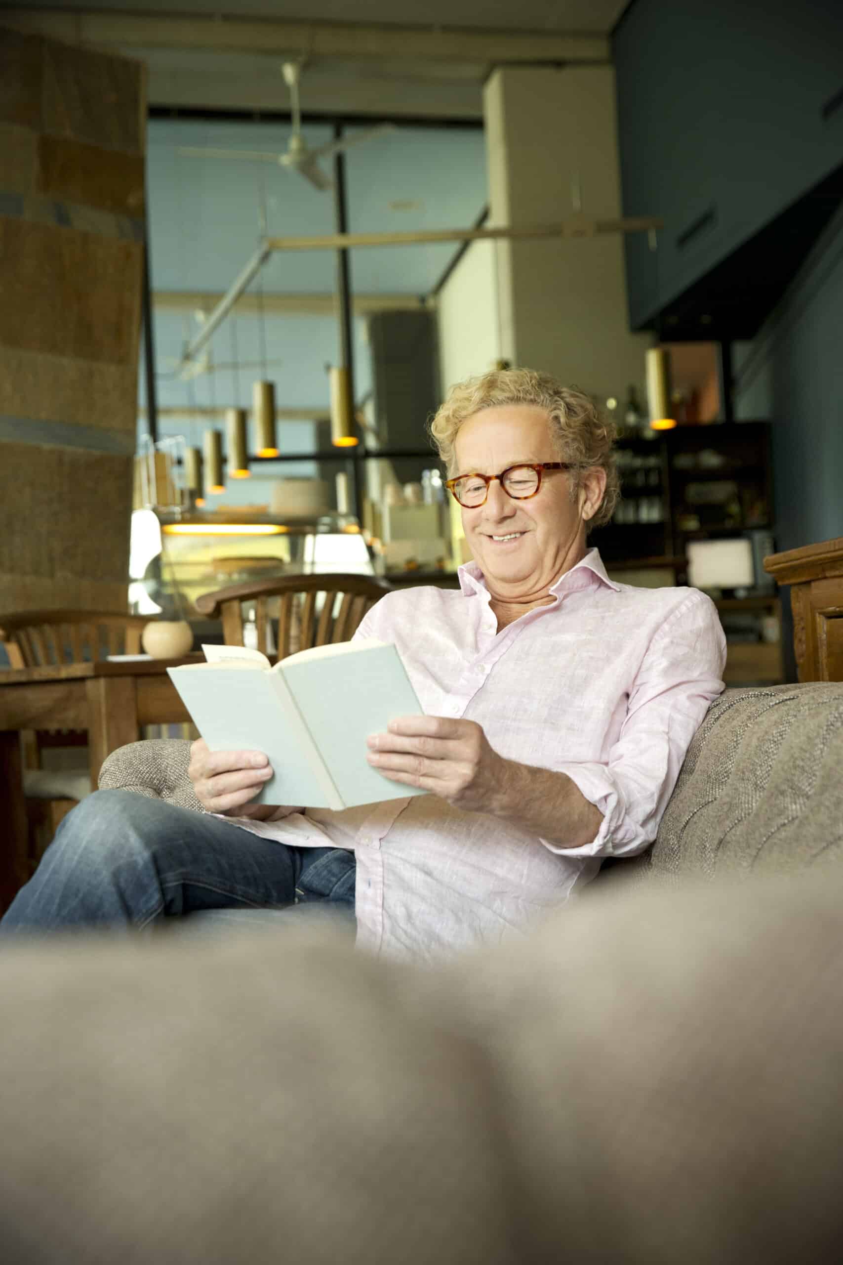 Ein Mann mit lockigem Haar und Brille, der ein hellrosa Hemd und Jeans trägt, sitzt gemütlich auf einem Sofa und liest ein Buch. Der Hintergrund zeigt ein modernes, gut beleuchtetes Interieur mit Hängelampen und Holzmöbeln. Er wirkt entspannt und lächelt. © Fotografie Tomas Rodriguez