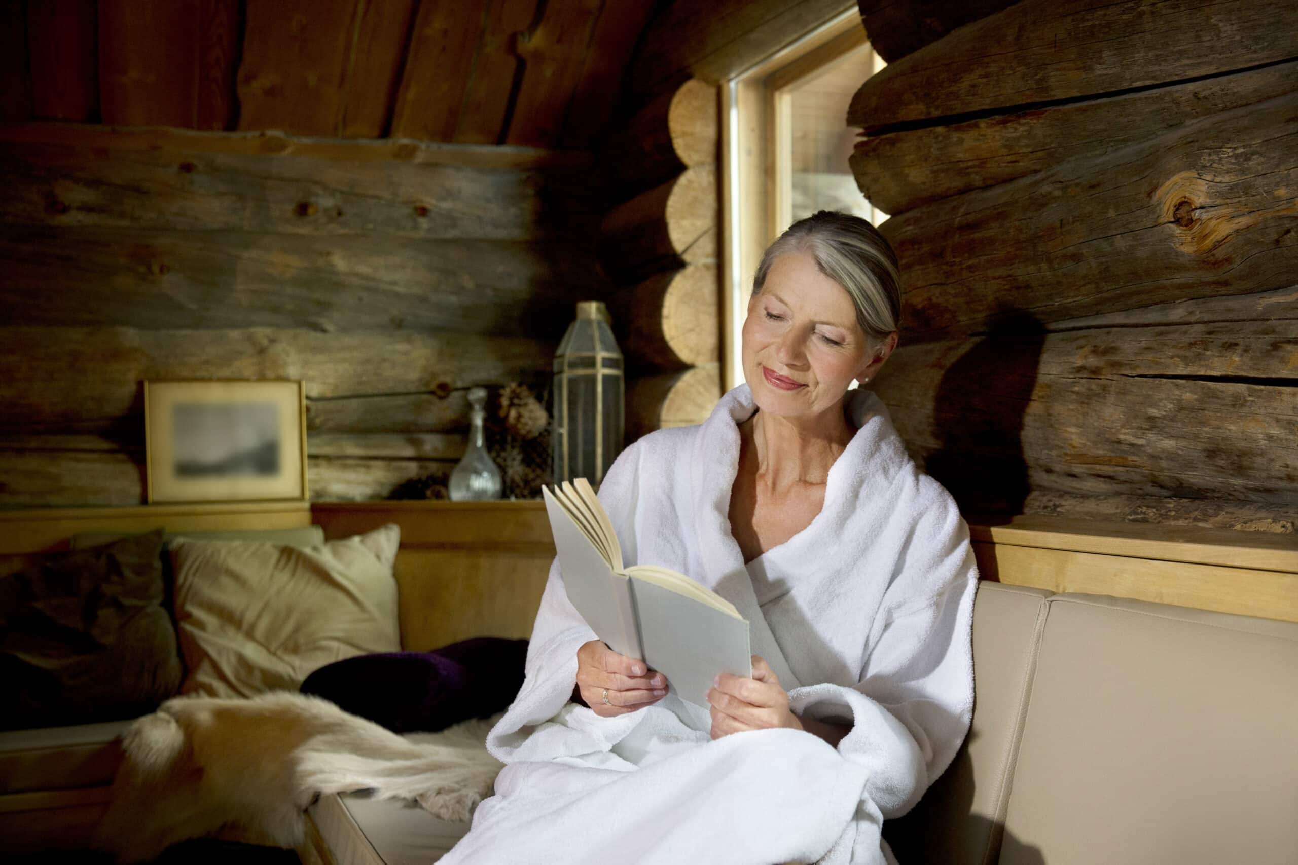 Eine Frau im weißen Bademantel sitzt auf einem beigen Sofa in einer rustikalen Blockhütte. Sie liest ein Buch und lächelt. Durch ein nahes Fenster fällt Tageslicht ein und erhellt die gemütliche Atmosphäre, die mit gerahmten Bildern und Dekoartikeln geschmückt ist. © Fotografie Tomas Rodriguez
