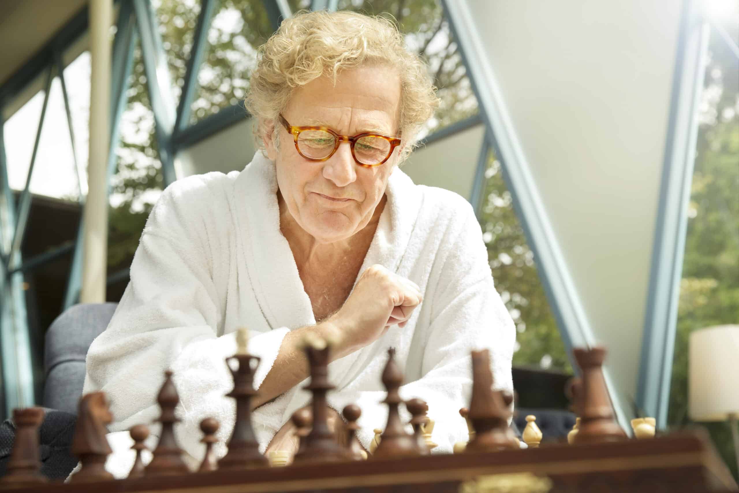 Ein Mann mit blondem Lockenhaar, Brille und weißem Bademantel sitzt nachdenklich vor einem Schachbrett. Er scheint sich auf das Spiel zu konzentrieren. Sonnenlicht fällt durch die großen Fenster im Hintergrund und erhellt die Szene. © Fotografie Tomas Rodriguez