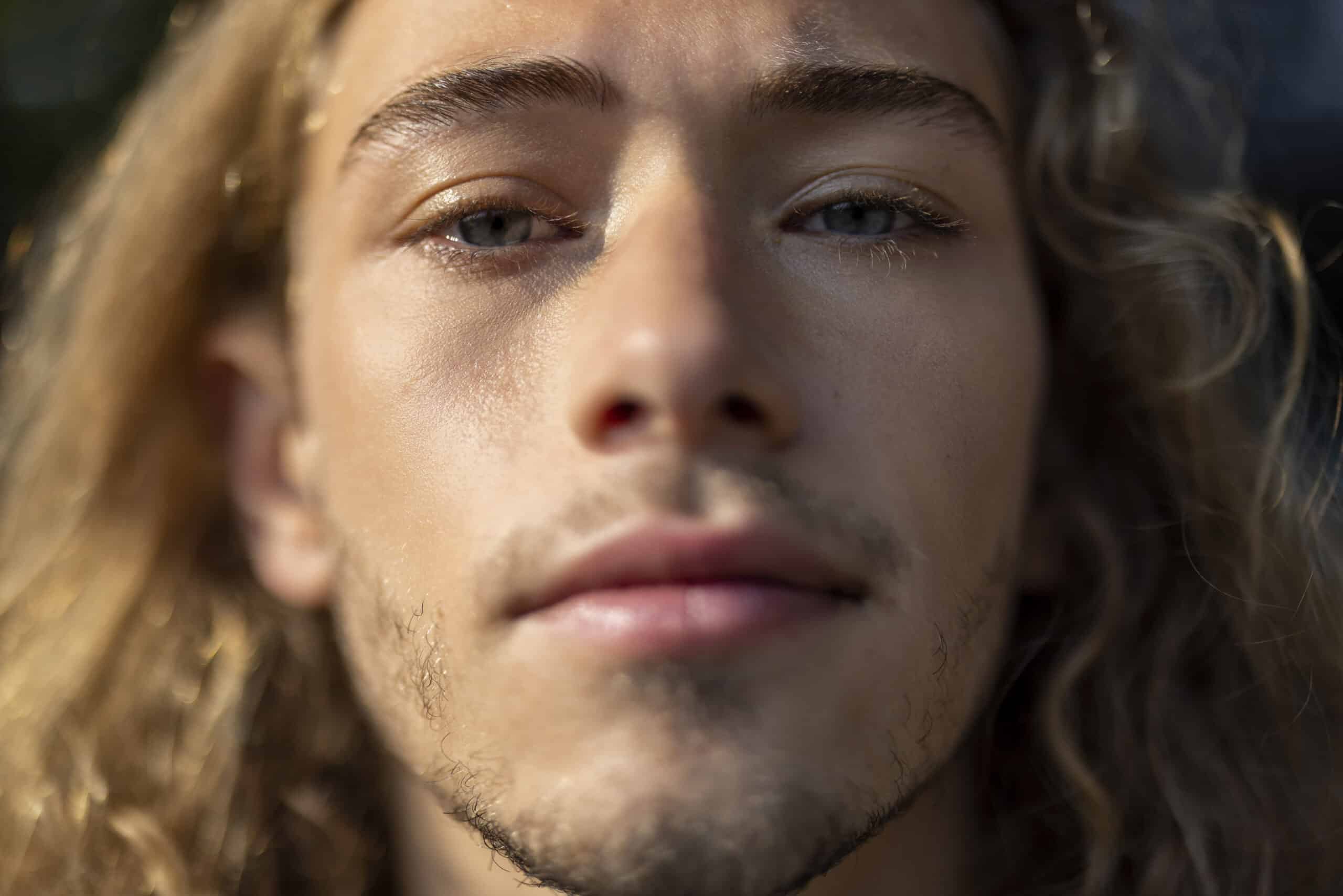 Nahaufnahme einer Person mit langen, welligen blonden Haaren und hellen Augen, die direkt in die Kamera blickt. Der Hintergrund ist unscharf, sodass die Aufmerksamkeit auf das Gesicht der Person gerichtet ist. Sie hat einen leichten Bart und einen neutralen Gesichtsausdruck. © Fotografie Tomas Rodriguez