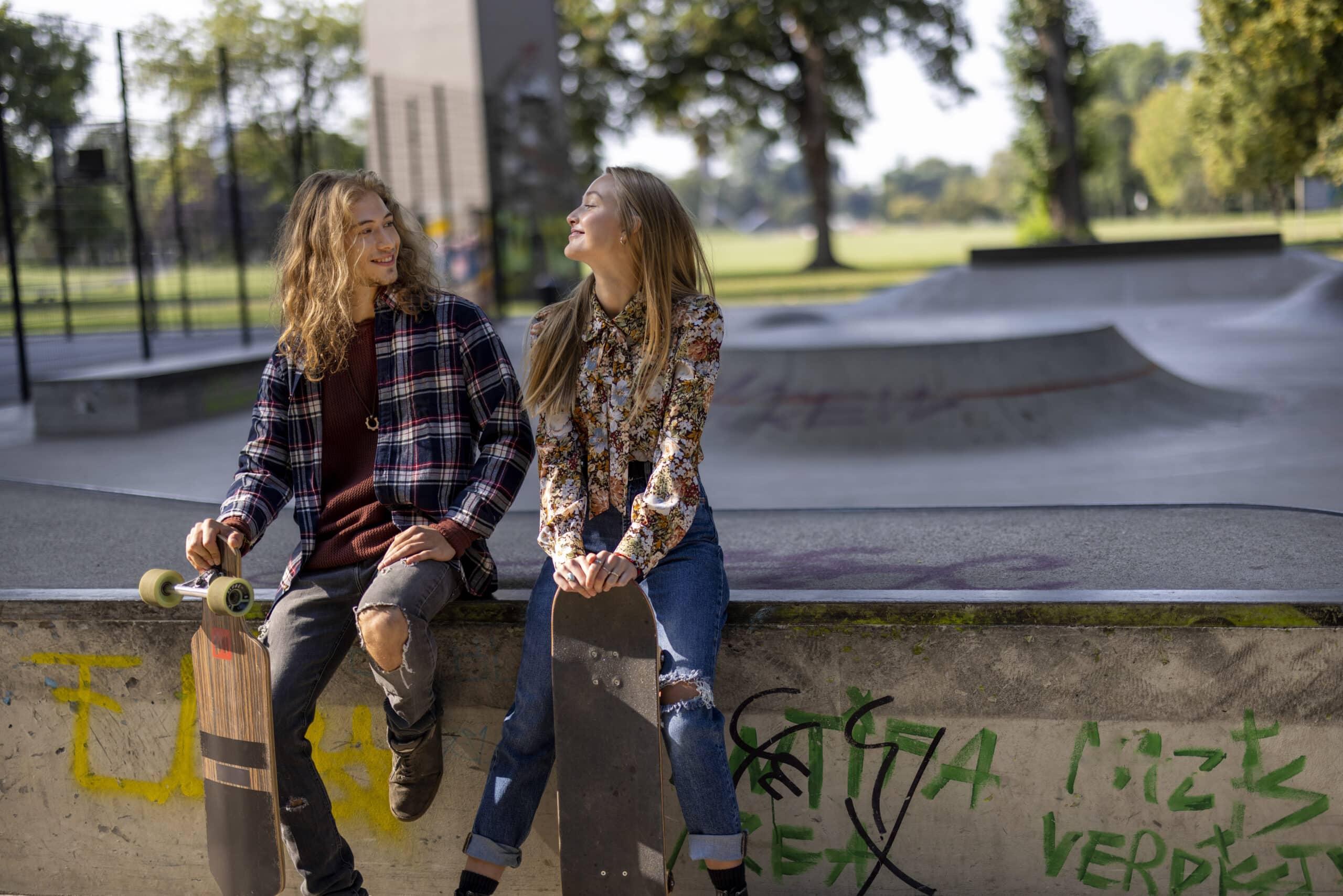 Zwei junge Leute, einer mit langen Haaren, trägt ein kariertes Hemd und hält ein Skateboard, der andere mit langen blonden Haaren, trägt ein geblümtes Hemd und hält ein Skateboard, sitzen am Rand eines mit Graffiti bedeckten Skatepark-Vorsprungs. Sie lächeln sich an. Im Hintergrund sind Bäume und Grünflächen zu sehen. © Fotografie Tomas Rodriguez