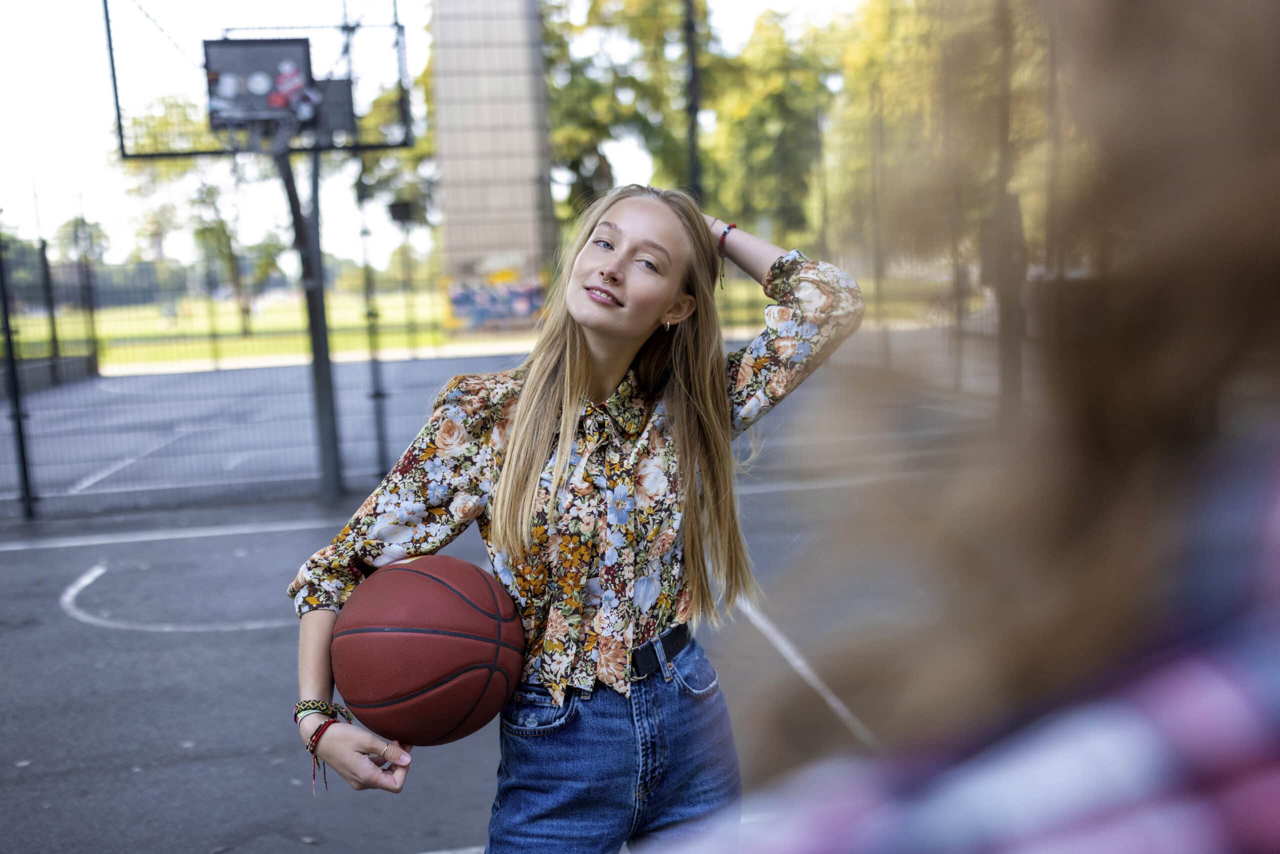 Eine junge Person mit langen Haaren steht auf einem Basketballplatz, hält einen Basketball unter einem Arm und legt die andere Hand auf den Kopf. Sie trägt ein Langarmshirt mit Blumenmuster und Jeans und lächelt in die Kamera. Im Vordergrund ist eine weitere Person unscharf zu sehen. © Fotografie Tomas Rodriguez