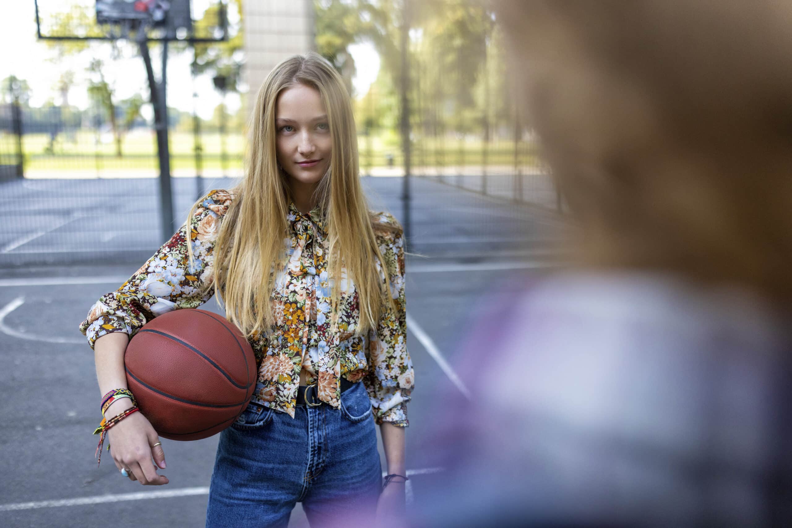 Eine junge Frau mit langen blonden Haaren, einer geblümten Bluse und Jeans steht auf einem Basketballplatz im Freien und hält einen Basketball unter dem Arm. Sie blickt selbstbewusst zu einer anderen Person, die der Kamera den Rücken zuwendet. Im Hintergrund ist ein Basketballkorb zu sehen. © Fotografie Tomas Rodriguez