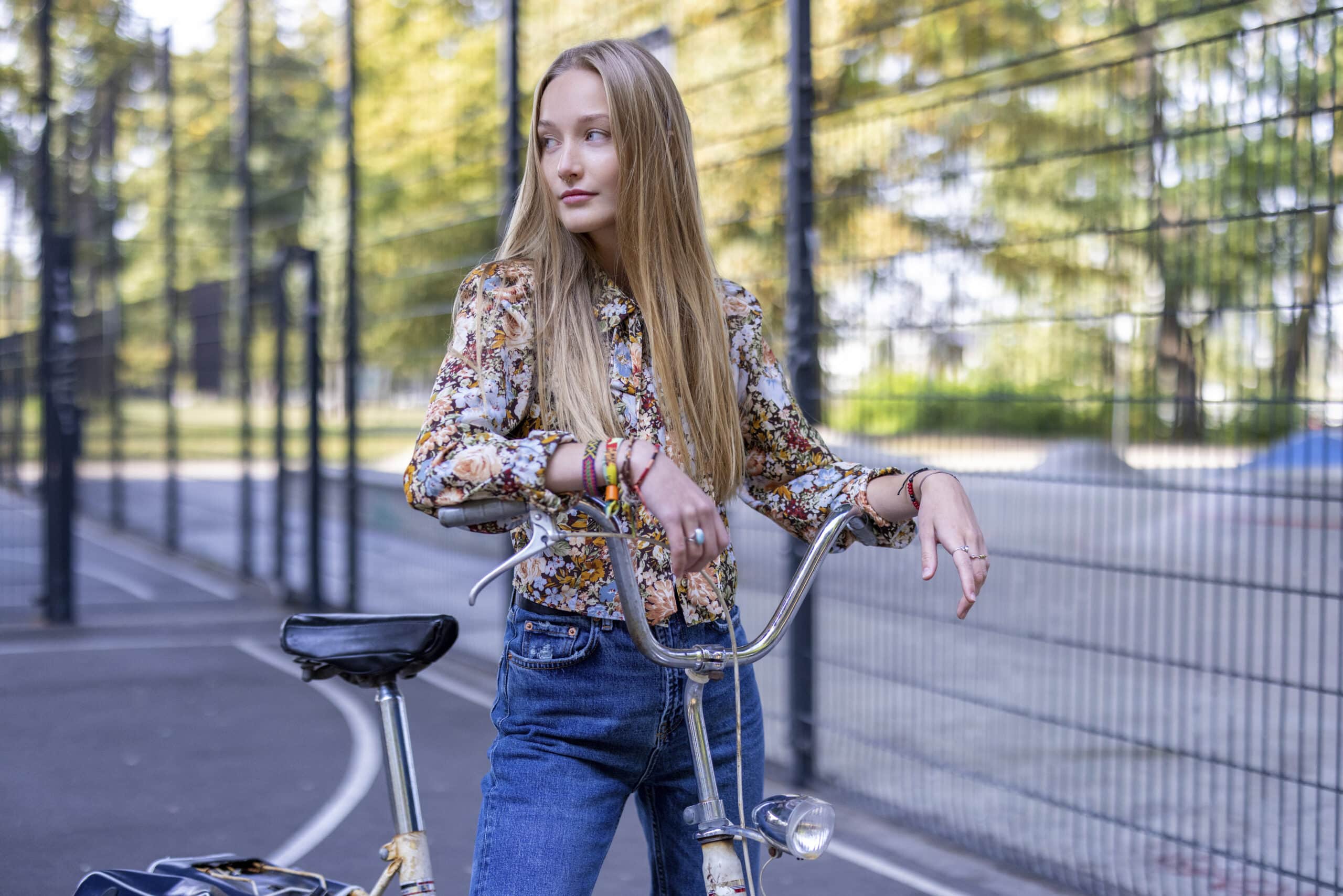 Eine junge Frau mit langen blonden Haaren, einer geblümten Bluse und Bluejeans, lehnt auf einem Vintage-Fahrrad. Sie steht in der Nähe eines eingezäunten Bereichs mit Bäumen und einem Park im Hintergrund und blickt mit nachdenklichem Gesichtsausdruck zur Seite. © Fotografie Tomas Rodriguez