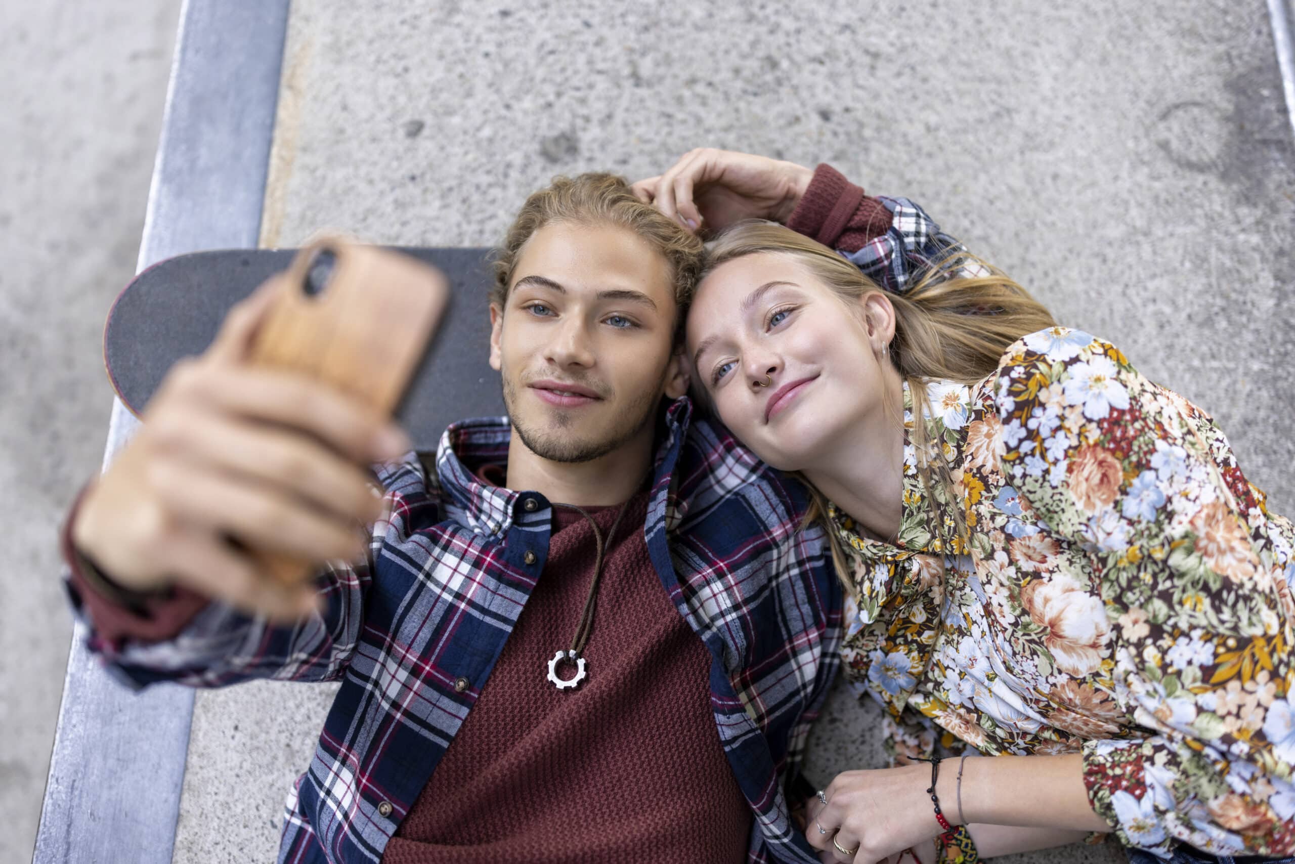 Ein junges Paar liegt eng beieinander auf dem Bürgersteig. Der junge Mann trägt ein rotes Hemd unter einer karierten Jacke und hält ein Smartphone für ein Selfie in der Hand. Die junge Frau, die eine geblümte Bluse trägt, legt ihren Kopf auf seine Schulter und lächelt zufrieden. Neben ihnen liegt ein Skateboard. © Fotografie Tomas Rodriguez