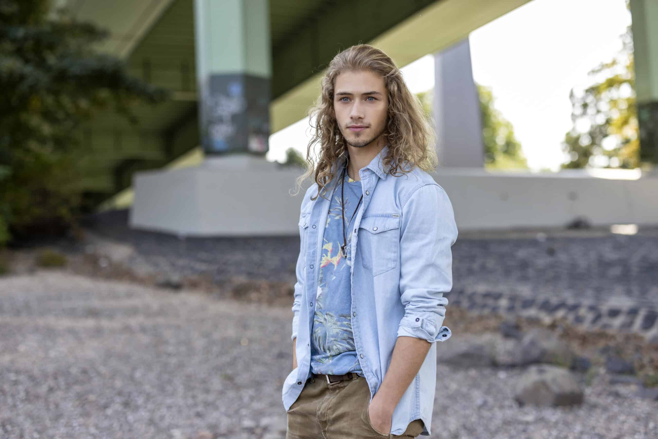 Ein junger Mann mit langem, welligem Haar steht unter einer Überführung. Er trägt ein helles Jeanshemd über einem bedruckten T-Shirt und hat die Hände in den Taschen seiner Khakihose. Im Hintergrund sind Säulen und Blätter zu sehen. Die Szenerie wirkt ungezwungen und im Freien. © Fotografie Tomas Rodriguez