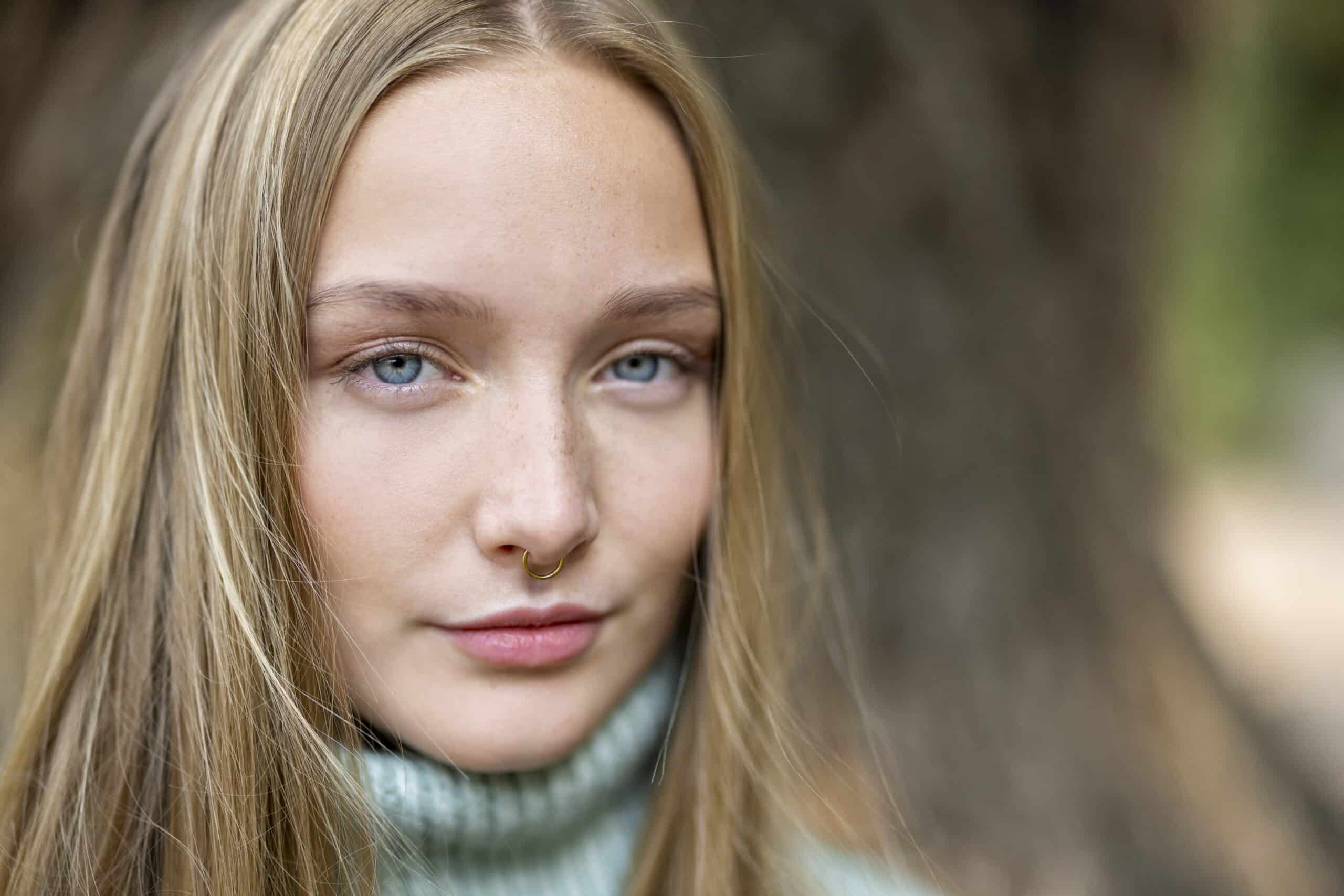 Eine junge Person mit langen blonden Haaren blickt direkt in die Kamera. Sie hat ein Septumpiercing und trägt einen gerippten Rollkragenpullover. Der Hintergrund ist unscharf und zeigt einen Baumstamm und Grünpflanzen. © Fotografie Tomas Rodriguez