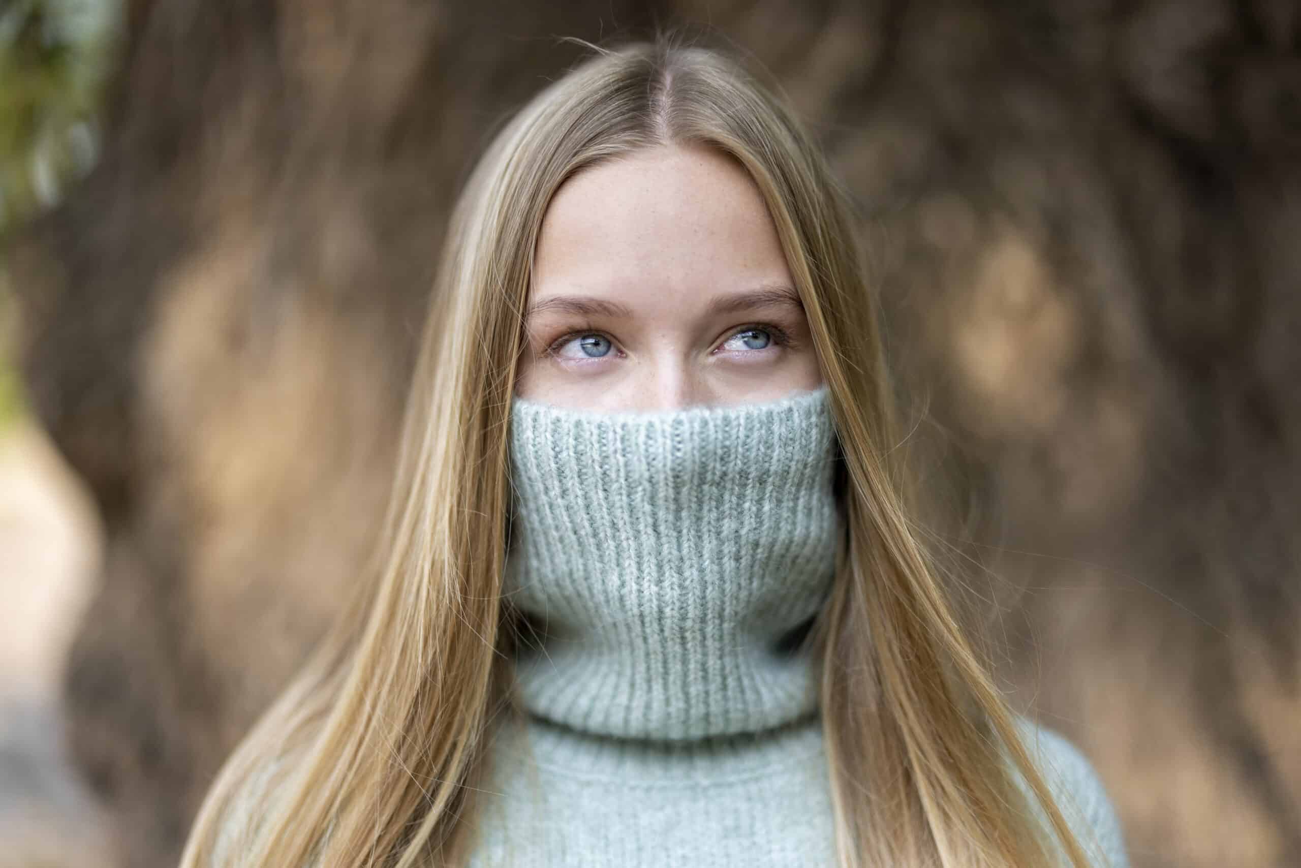 Eine junge Person mit langen blonden Haaren steht vor einem unscharfen natürlichen Hintergrund. Sie trägt einen hellgrauen Strickpullover und hat den hohen Kragen so hochgezogen, dass Mund und Nase bedeckt sind und nur die Augen frei bleiben. Sie blickt nachdenklich zur Seite. © Fotografie Tomas Rodriguez