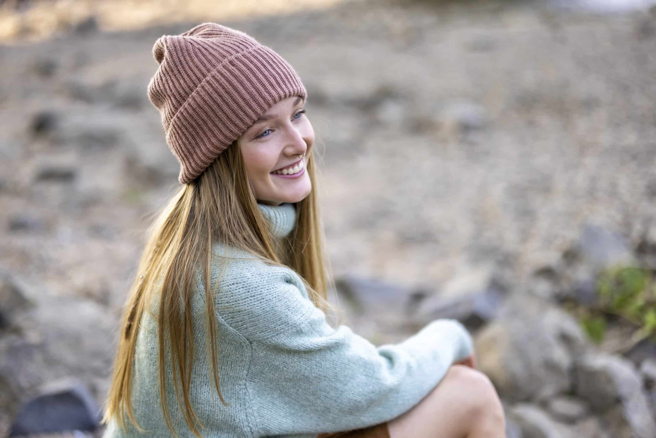 Eine Frau mit langen Haaren, einer braunen Strickmütze und einem hellgrünen Pullover sitzt draußen auf einem felsigen Untergrund. Sie blickt nach links und lächelt. Der Hintergrund ist leicht verschwommen, was die natürliche, ruhige Umgebung hervorhebt. © Fotografie Tomas Rodriguez