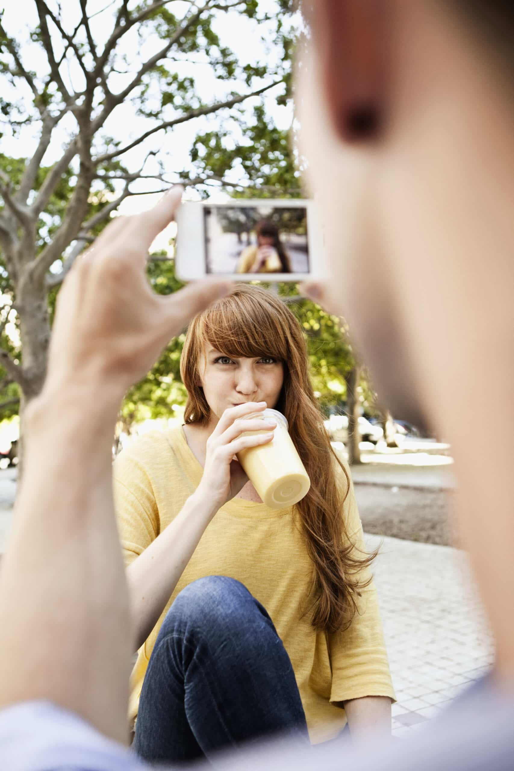 Eine Frau mit langen roten Haaren und einem gelben Hemd trinkt aus einer Flasche, während jemand mit einem Smartphone ein Foto von ihr macht. Sie sitzt im Freien mit Bäumen und Sonnenlicht im Hintergrund. © Fotografie Tomas Rodriguez